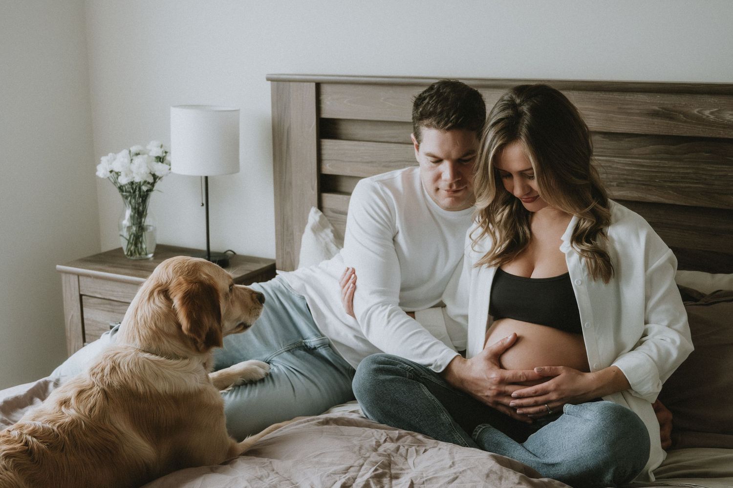 Pregnant couple sitting on bed with golden retriever dog in cozy bedroom.