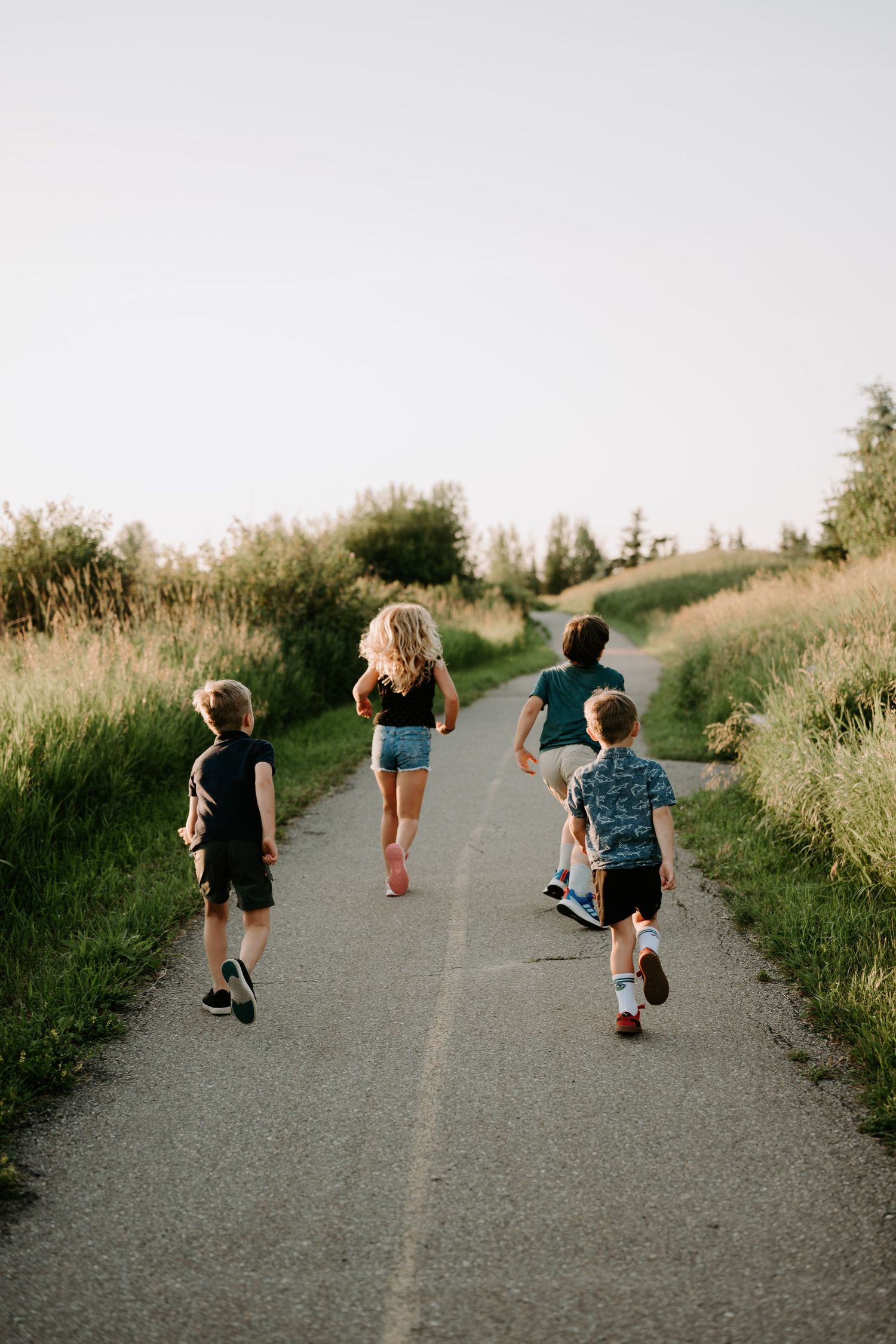 Four children walking away on a paved path surrounded by green grass and trees in warm evening light.