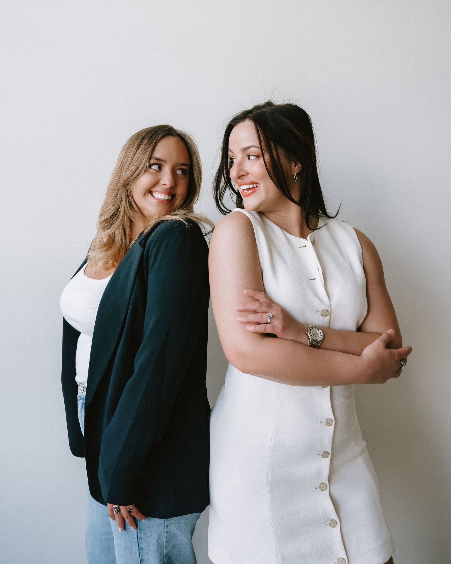 Two smiling women pose back-to-back, one in a black blazer and one in a white dress, against a light gray background.