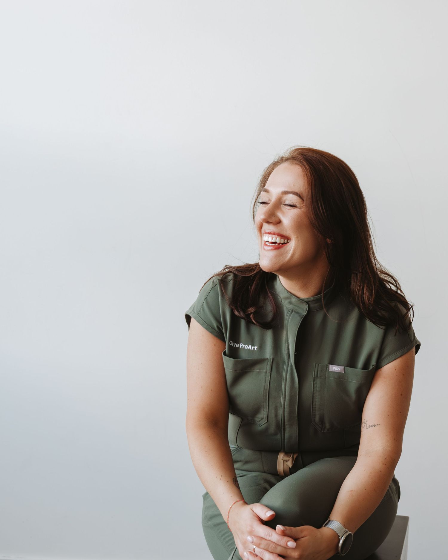 Woman in olive green jumpsuit laughing joyfully while seated against a light gray background.
