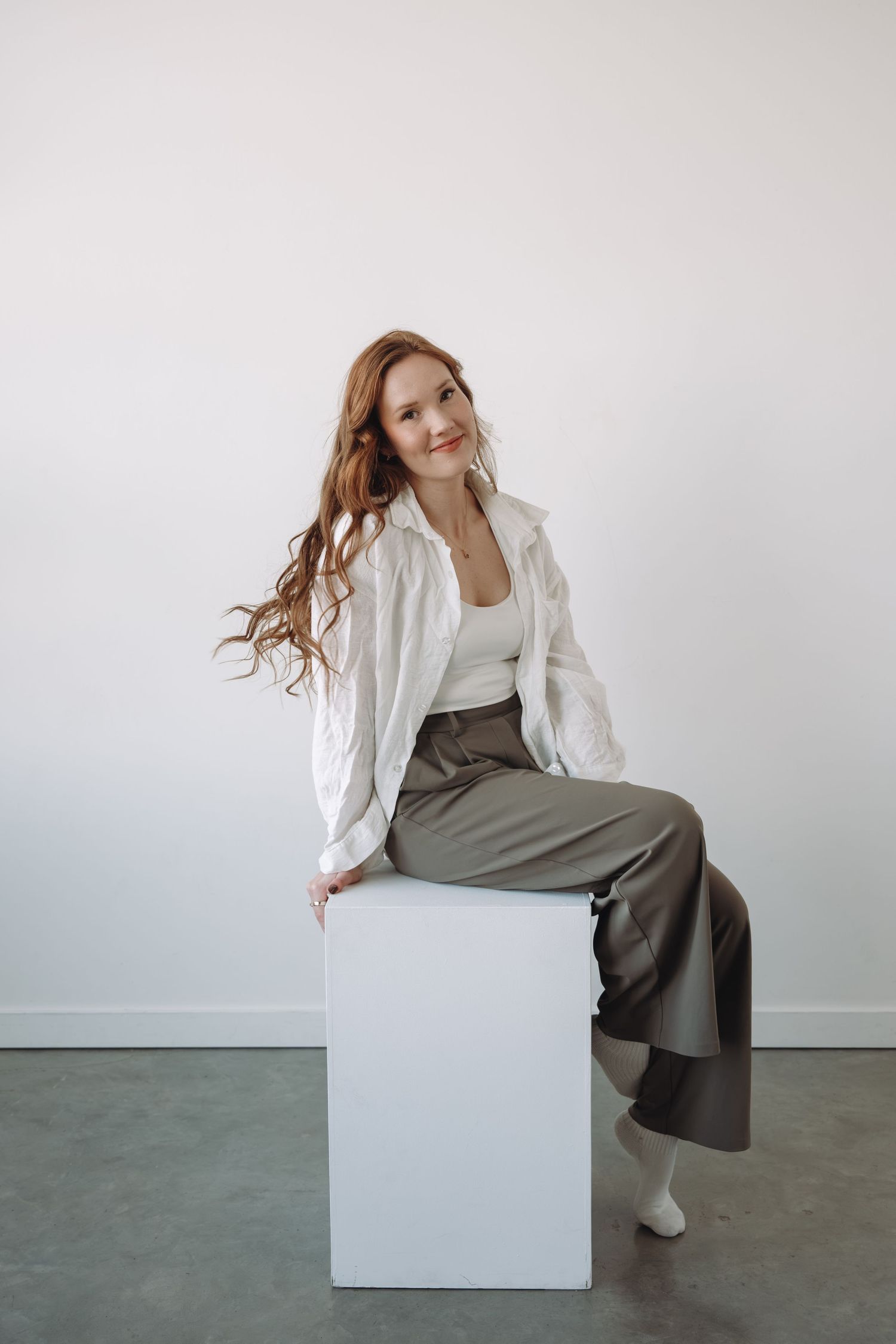 Smiling woman with long red hair in white jacket and olive pants sitting on white cube.