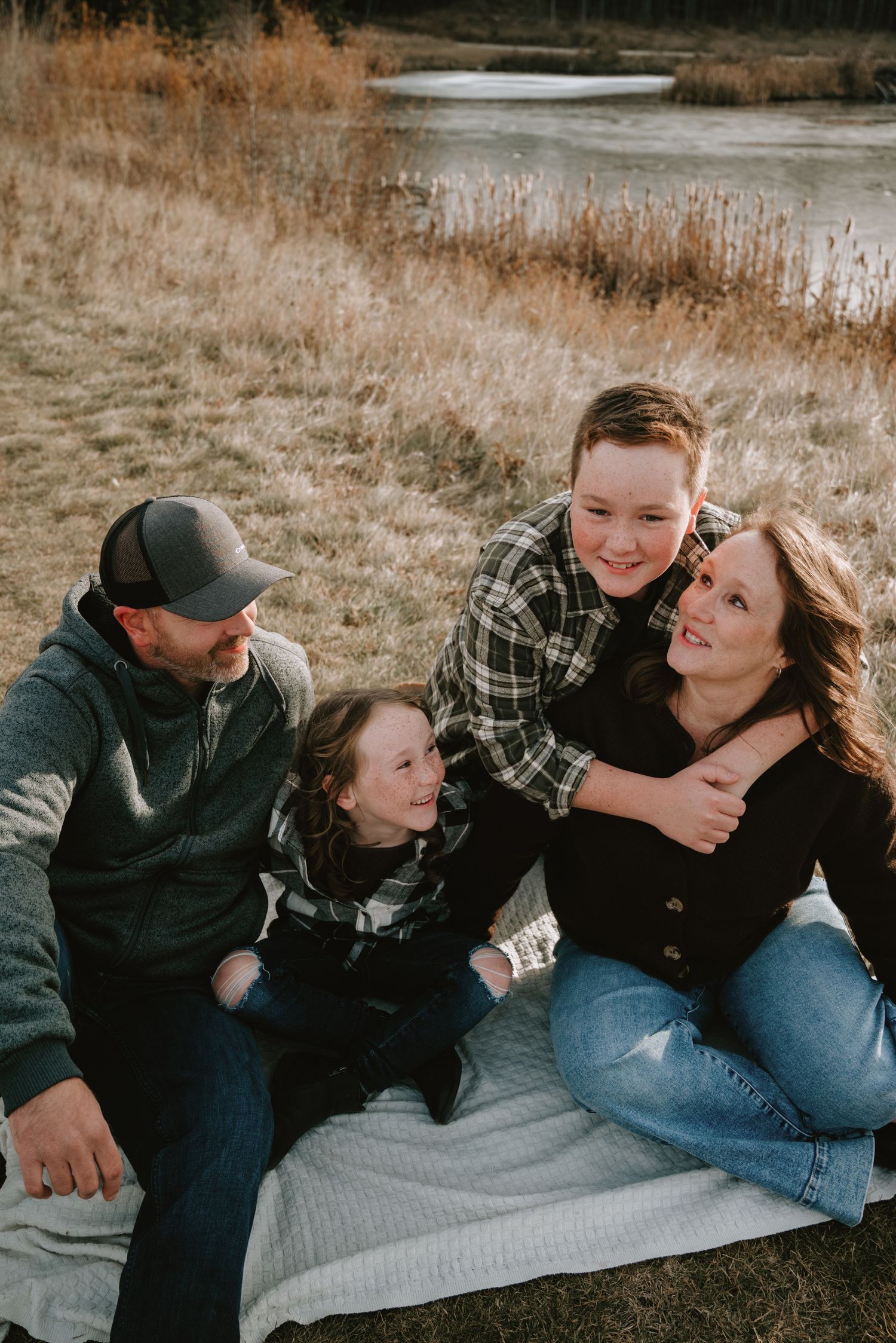 Happy family of four sitting on a blanket outdoors near a pond, laughing together during a fall photo session.