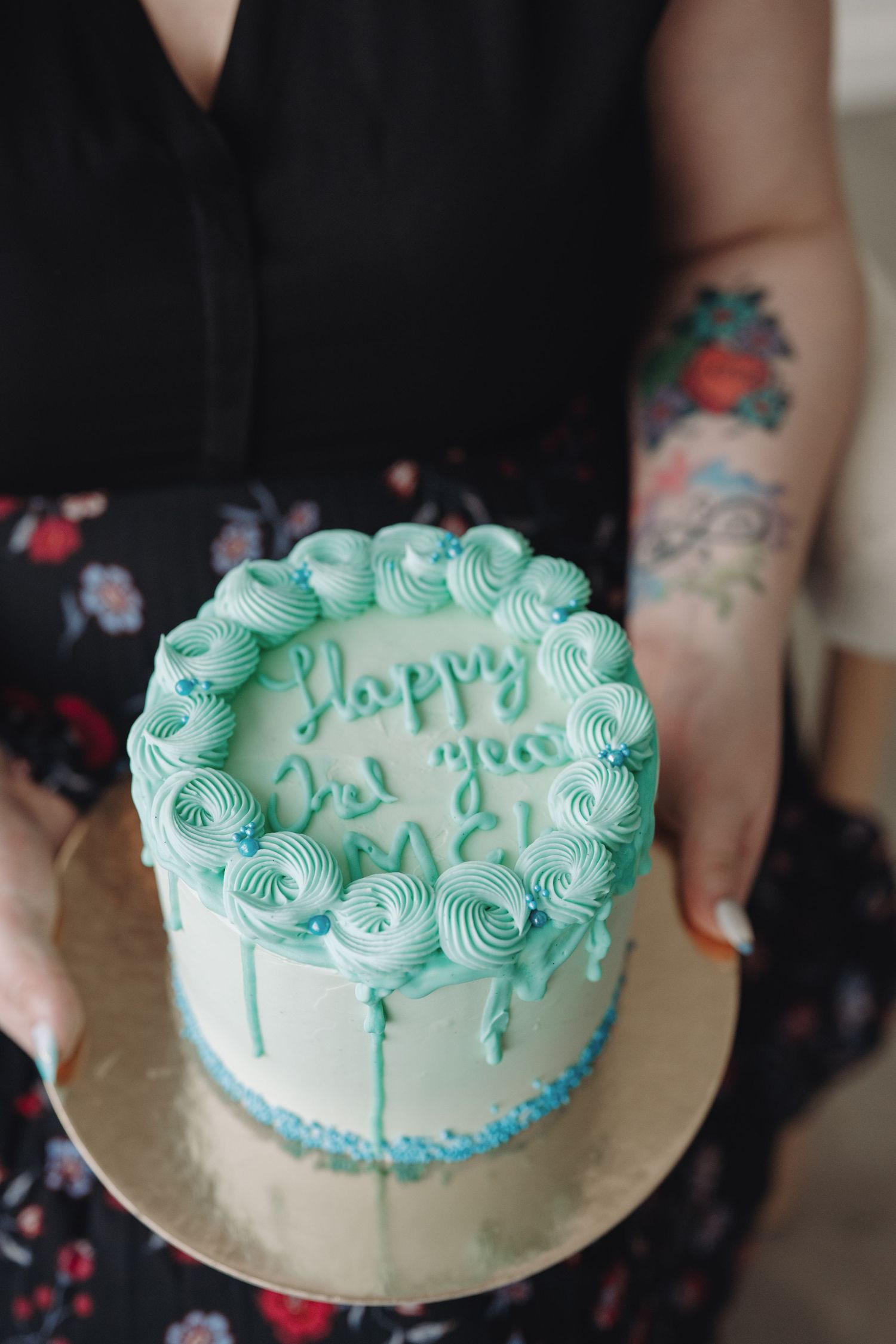 Person holding a teal frosted birthday cake with rosette decorations and Happy Birthday inscription on gold board.