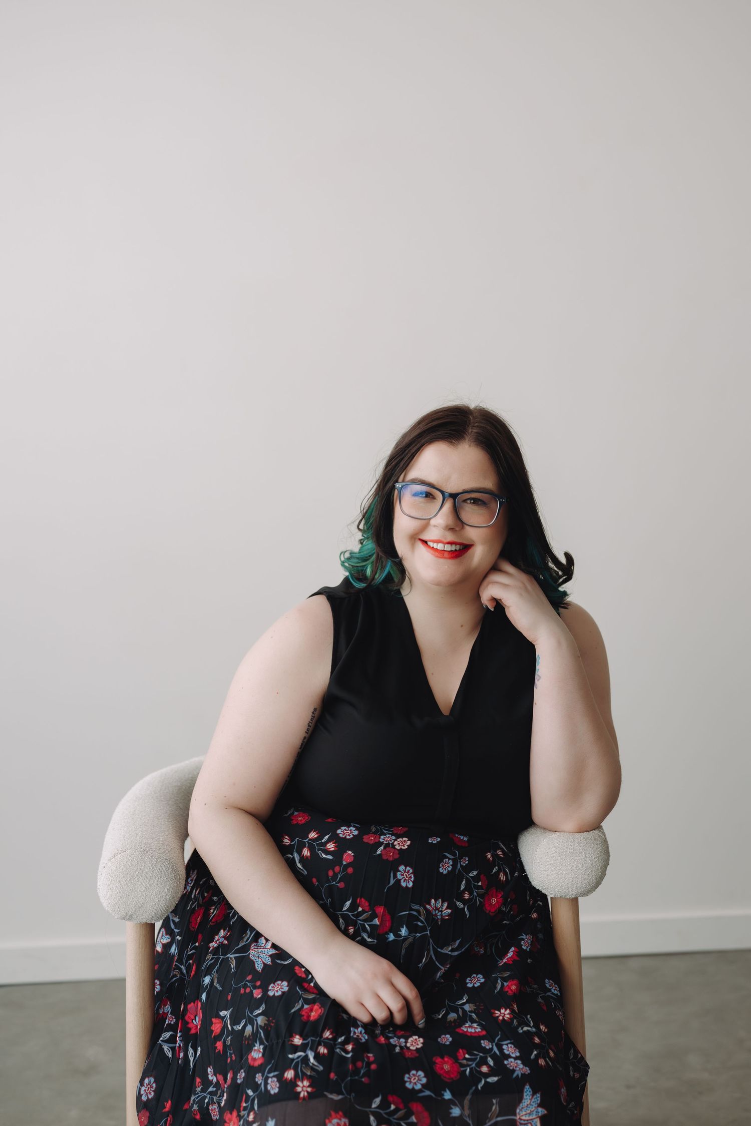 Woman with dark hair, glasses, and red lips sits smiling in a chair wearing a black floral dress.