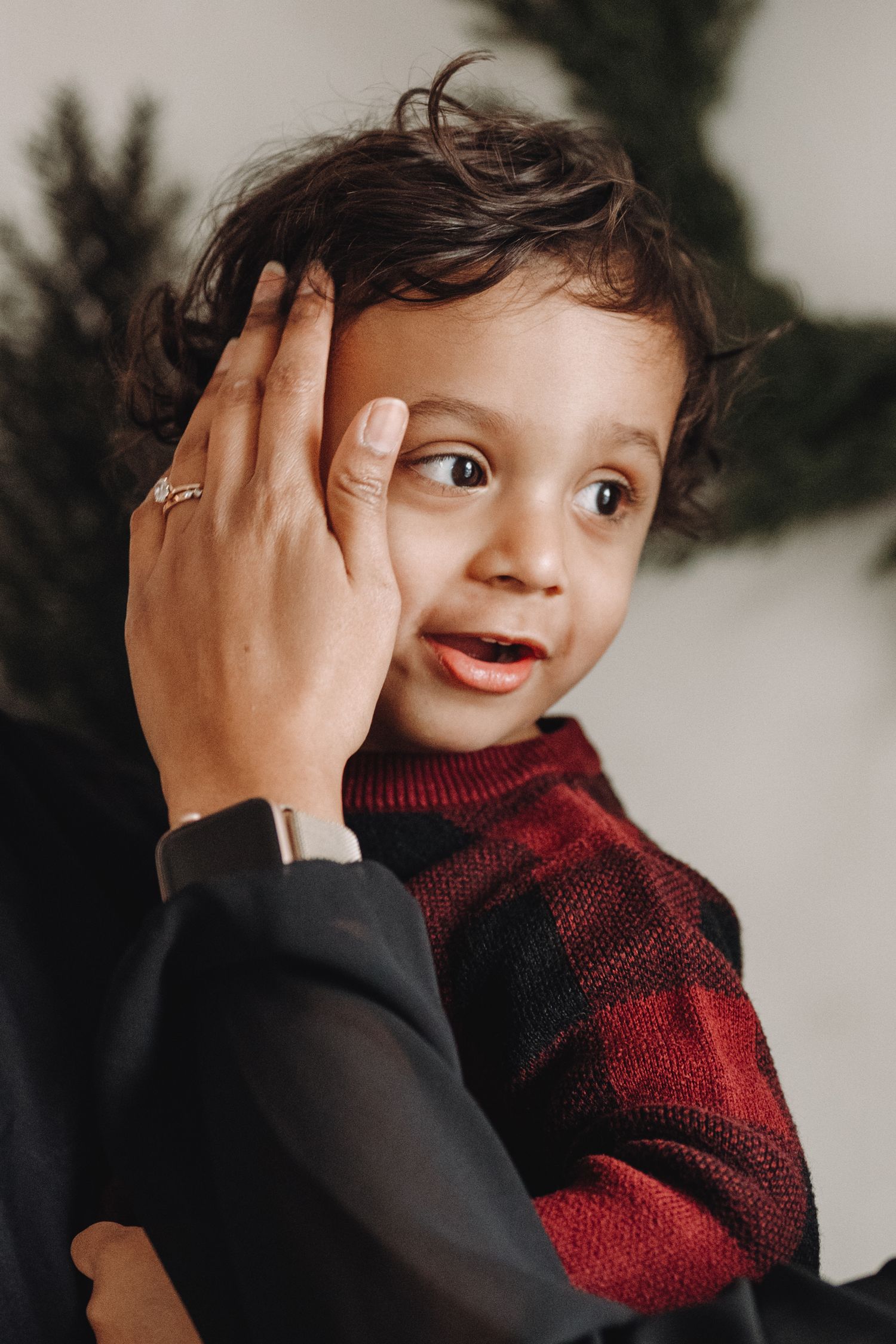 A curly-haired toddler in a red plaid shirt reacts expressively as an adult hand gently touches their face near a Christmas tree.