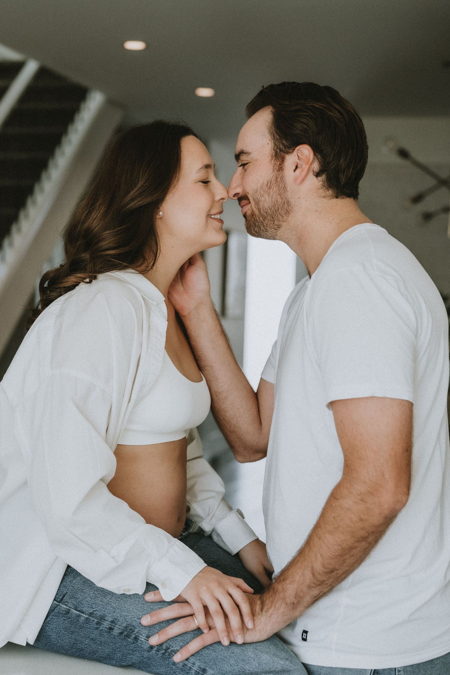A romantic couple in white tops share a tender moment, almost kissing, in a bright modern home interior.