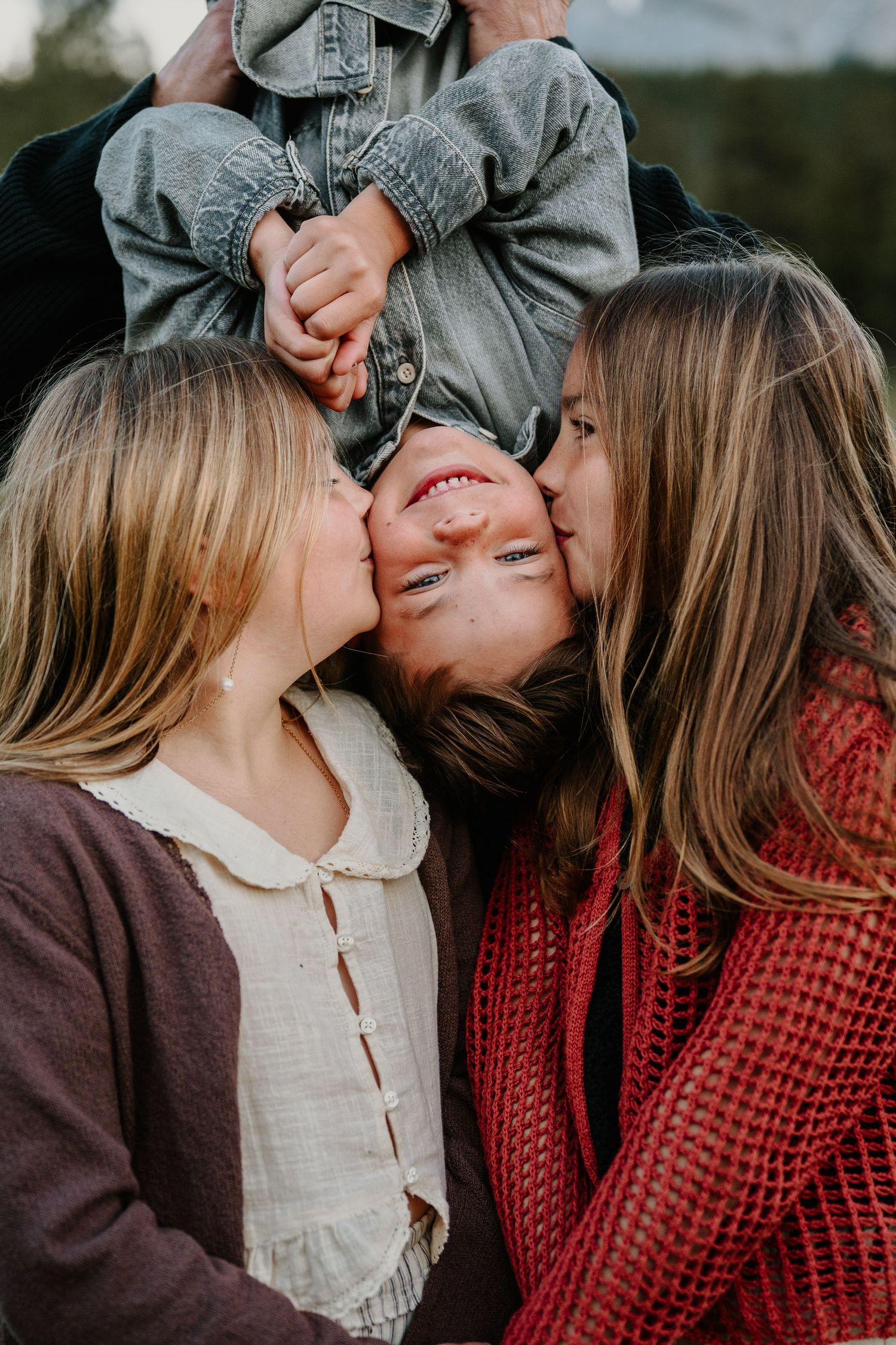Two girls kissing a laughing boy held upside down by an adult outdoors in a heartwarming family moment.