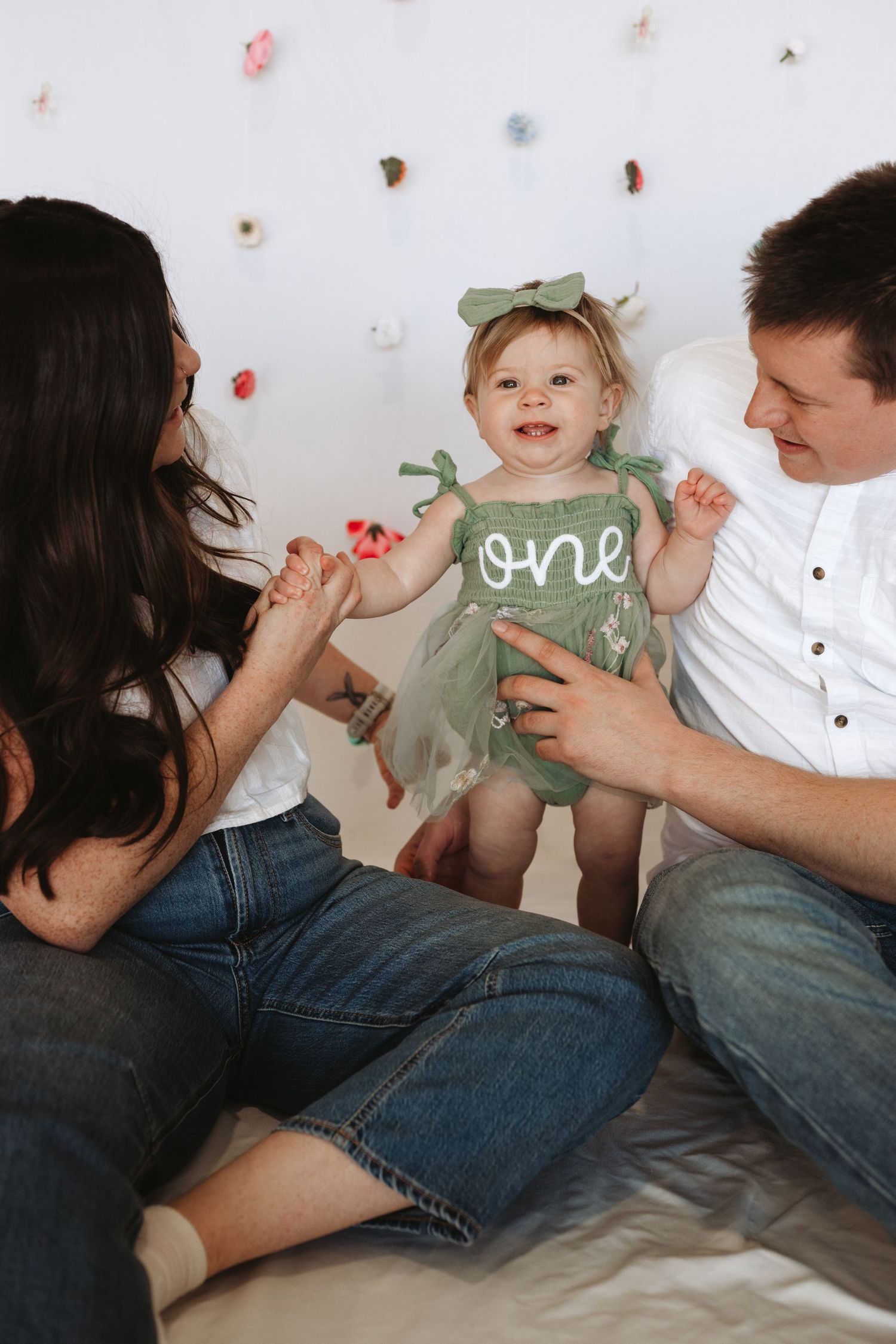 A baby girl in a green “one” tutu dress celebrates her first birthday with her smiling parents on a white backdrop.