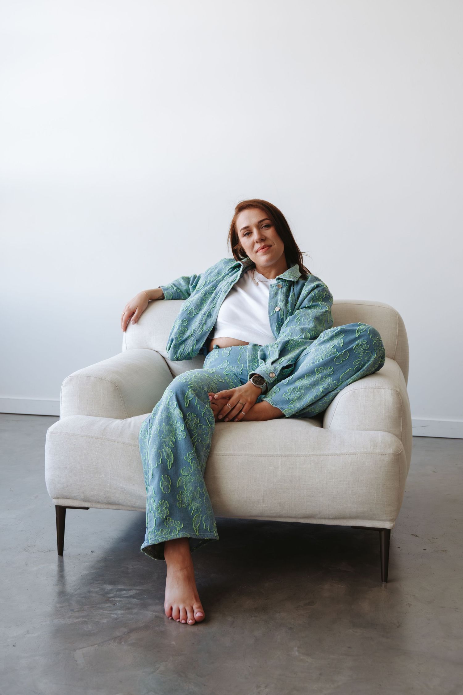 Woman in teal printed matching set relaxes on a cream armchair against a white wall on a concrete floor.
