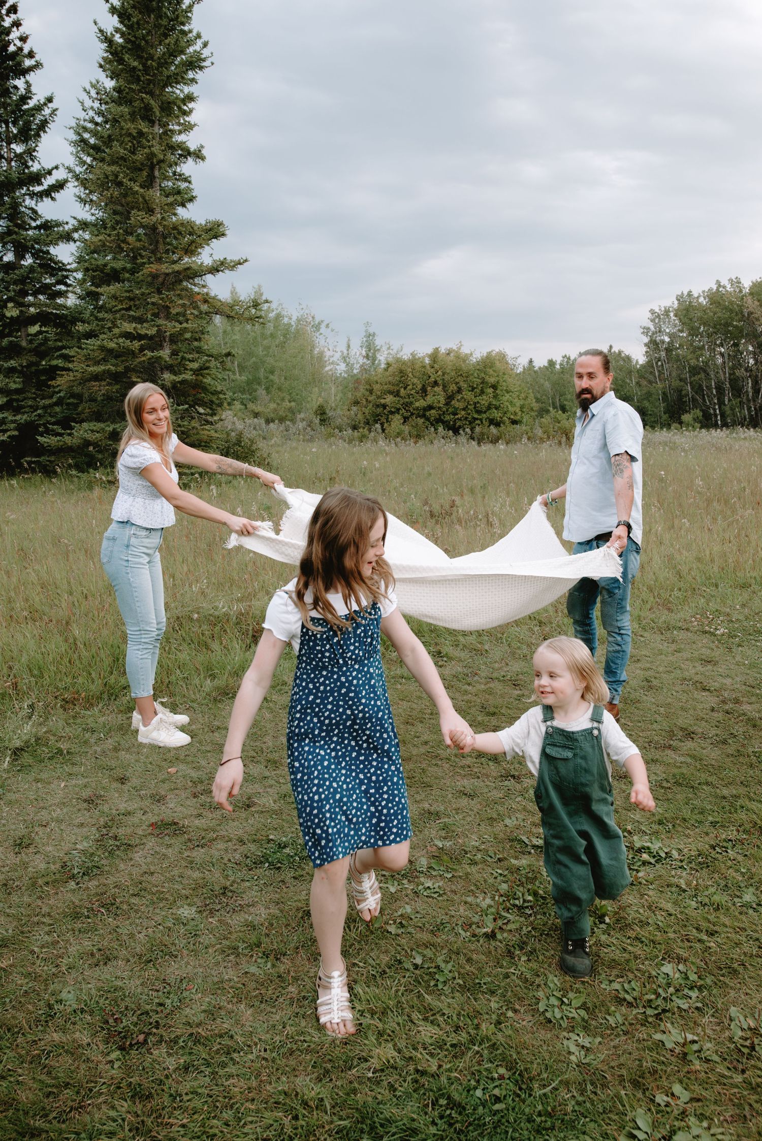 Happy family laughing and playing with white blanket in lush green meadow with pine trees in background.