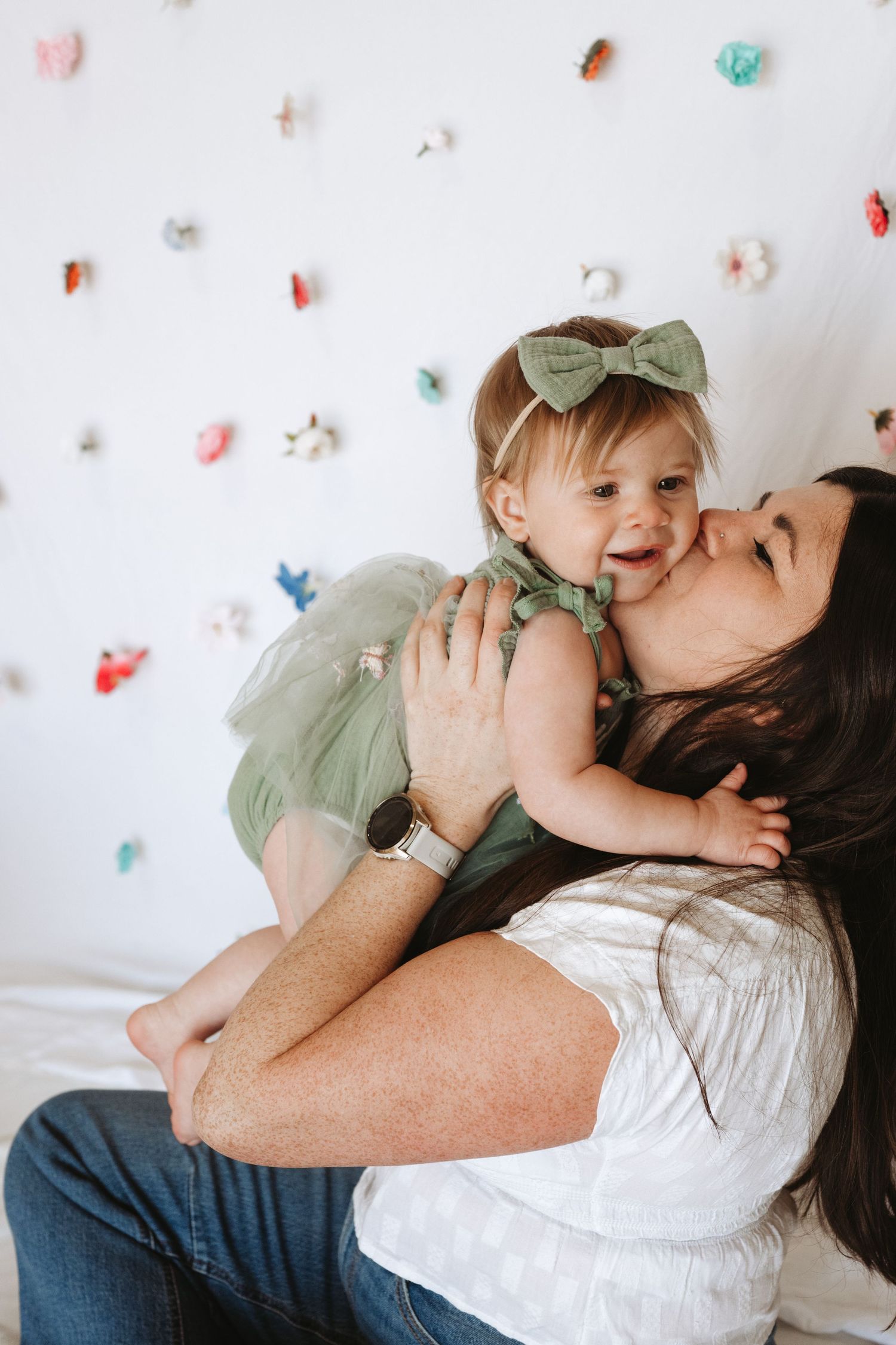 A mother kisses her smiling baby girl in a green tutu and bow headband against a colorful wall.