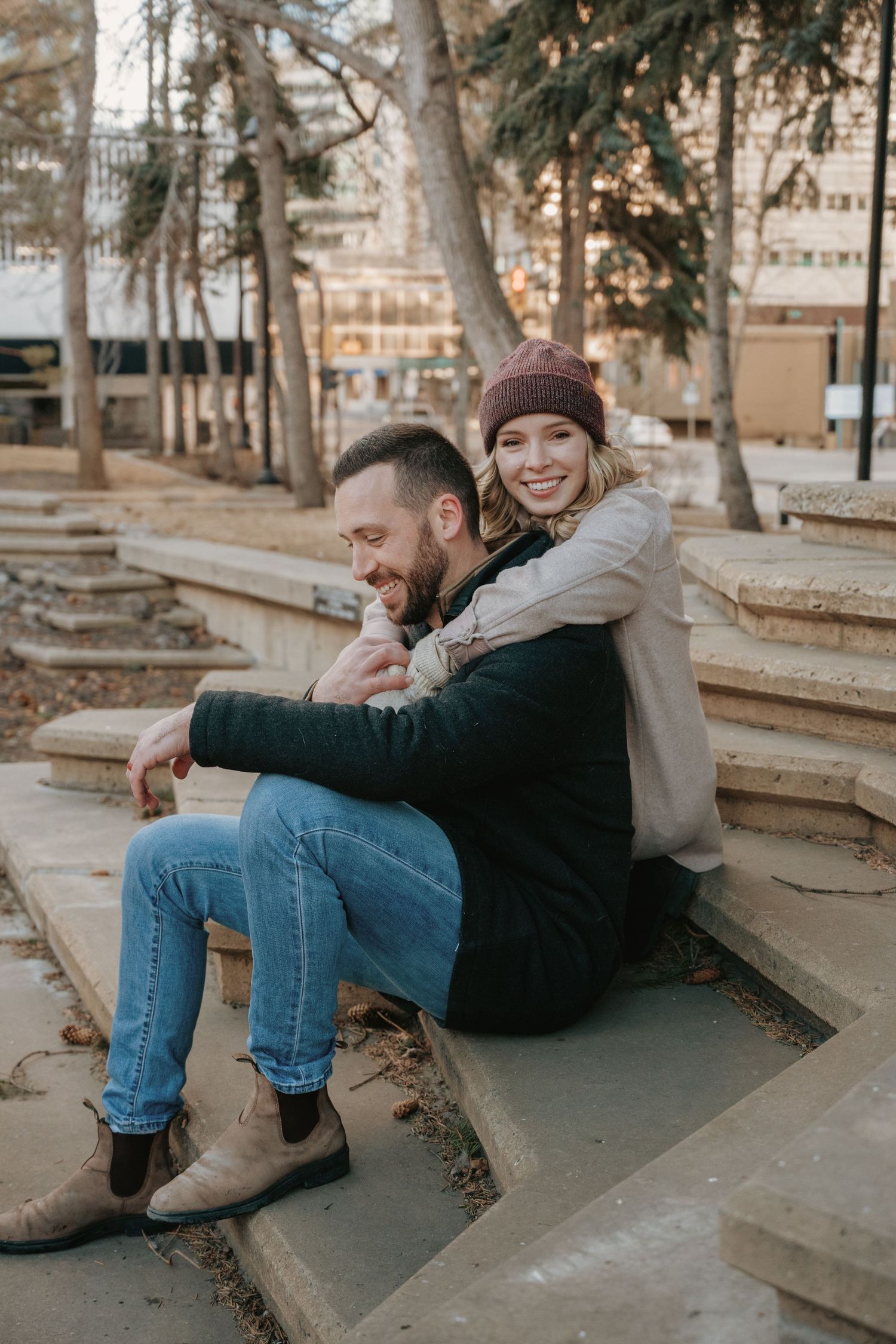 Couple sitting on outdoor steps in fall, woman in burgundy beanie hugging man in green jacket and jeans.