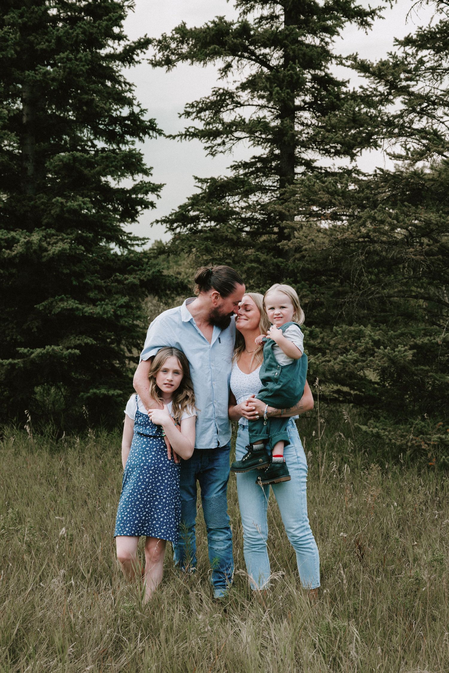 Family of four posing in a grassy field with evergreen trees, parents kissing while holding two young children.