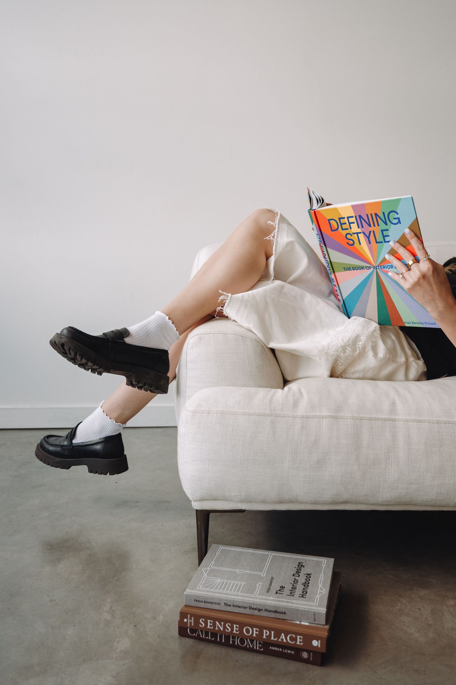 Person relaxing on white sofa reading colorful Defining Style book with stacked books on floor.