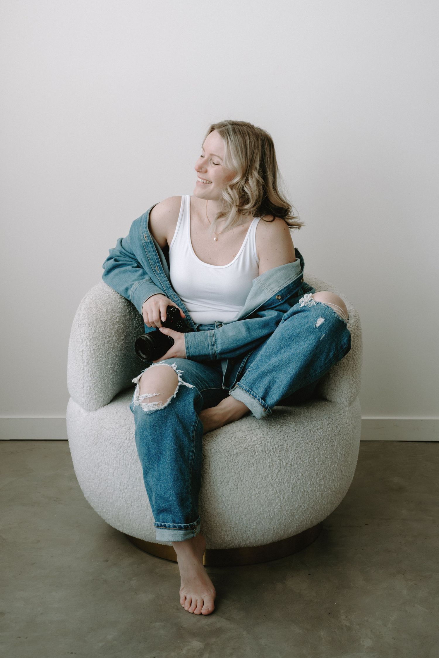 Smiling blonde woman relaxing in a round chair wearing jeans and white tank.