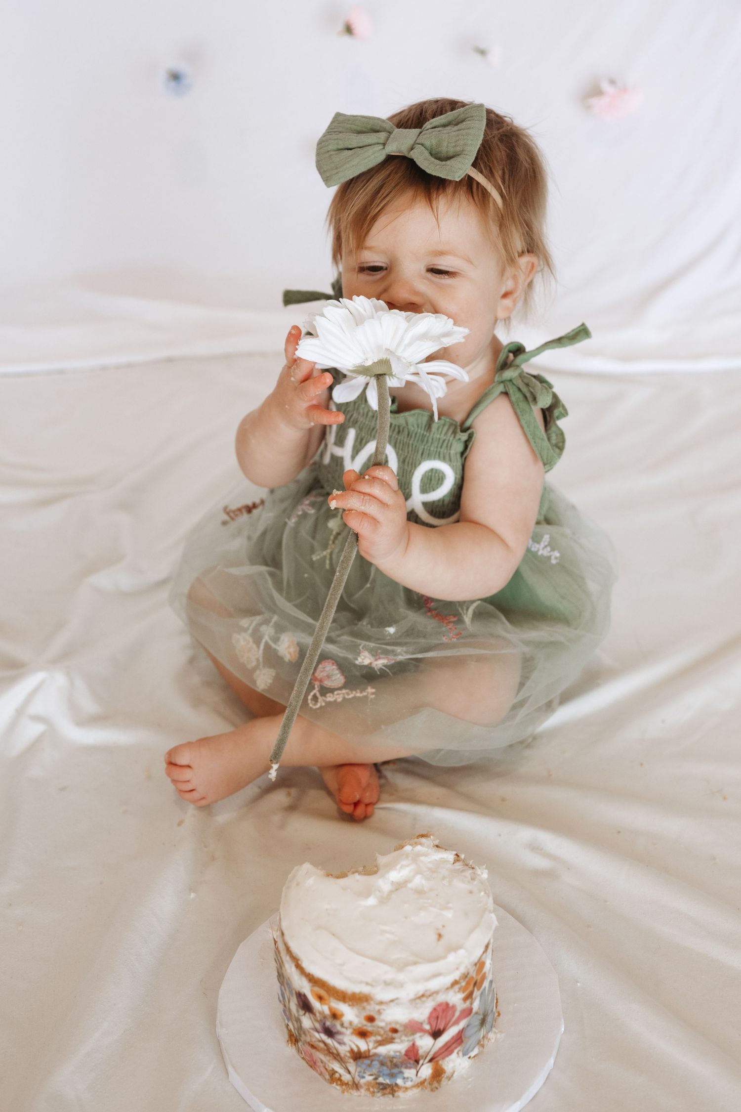 Baby girl in green tutu dress smells white daisy flower near smash cake on her first birthday.