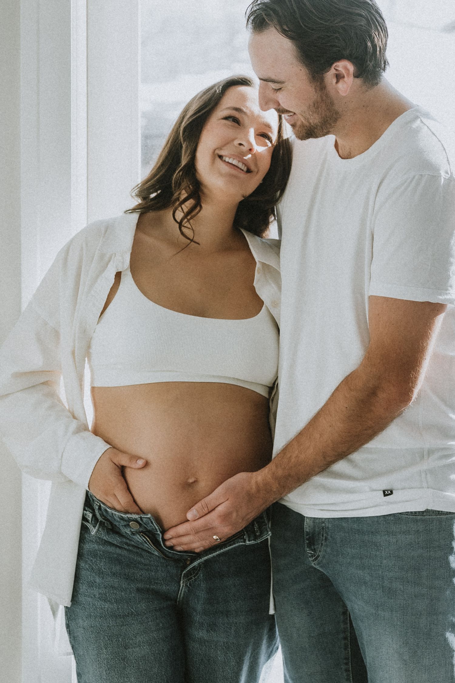 Happy pregnant couple in white tops and jeans sharing a tender moment while man gently touches baby bump.