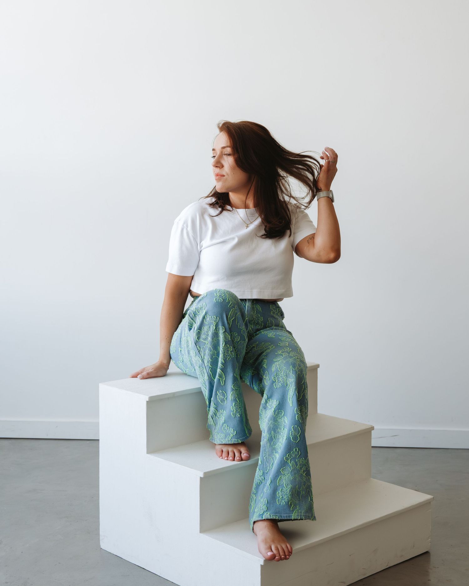 Brunette woman in white crop top and blue floral wide-leg pants sitting on white steps in bright studio.