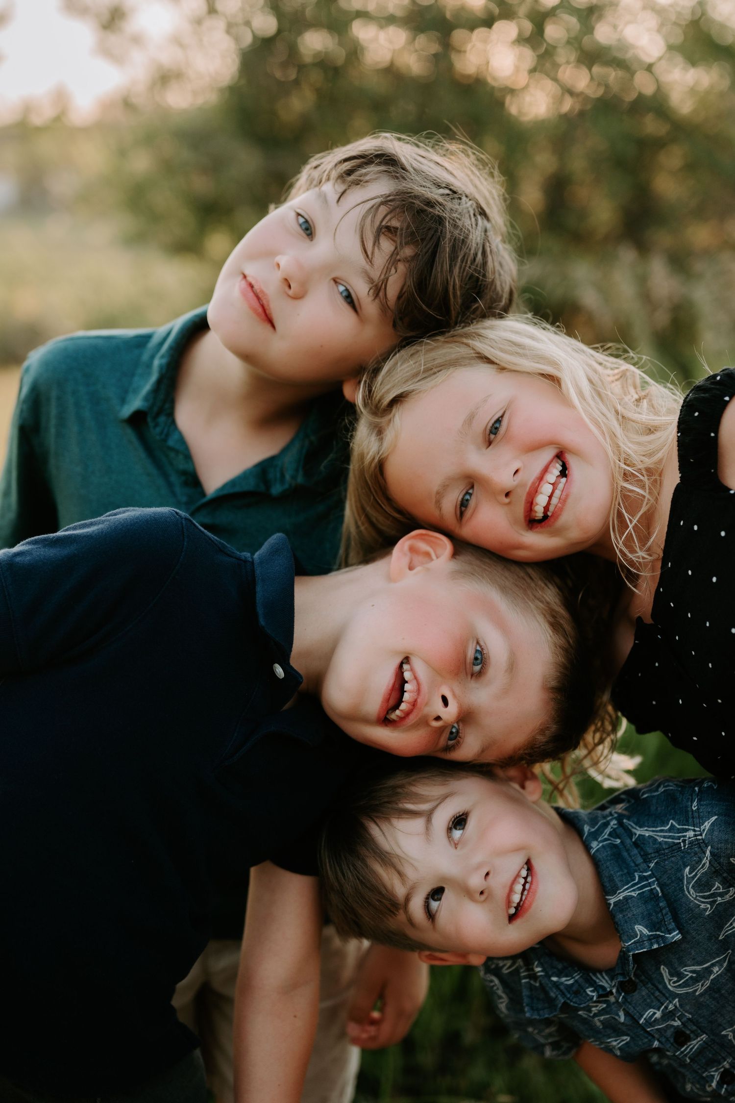Four smiling children huddle together outdoors in warm golden light, posing cheek to cheek in a joyful group portrait.