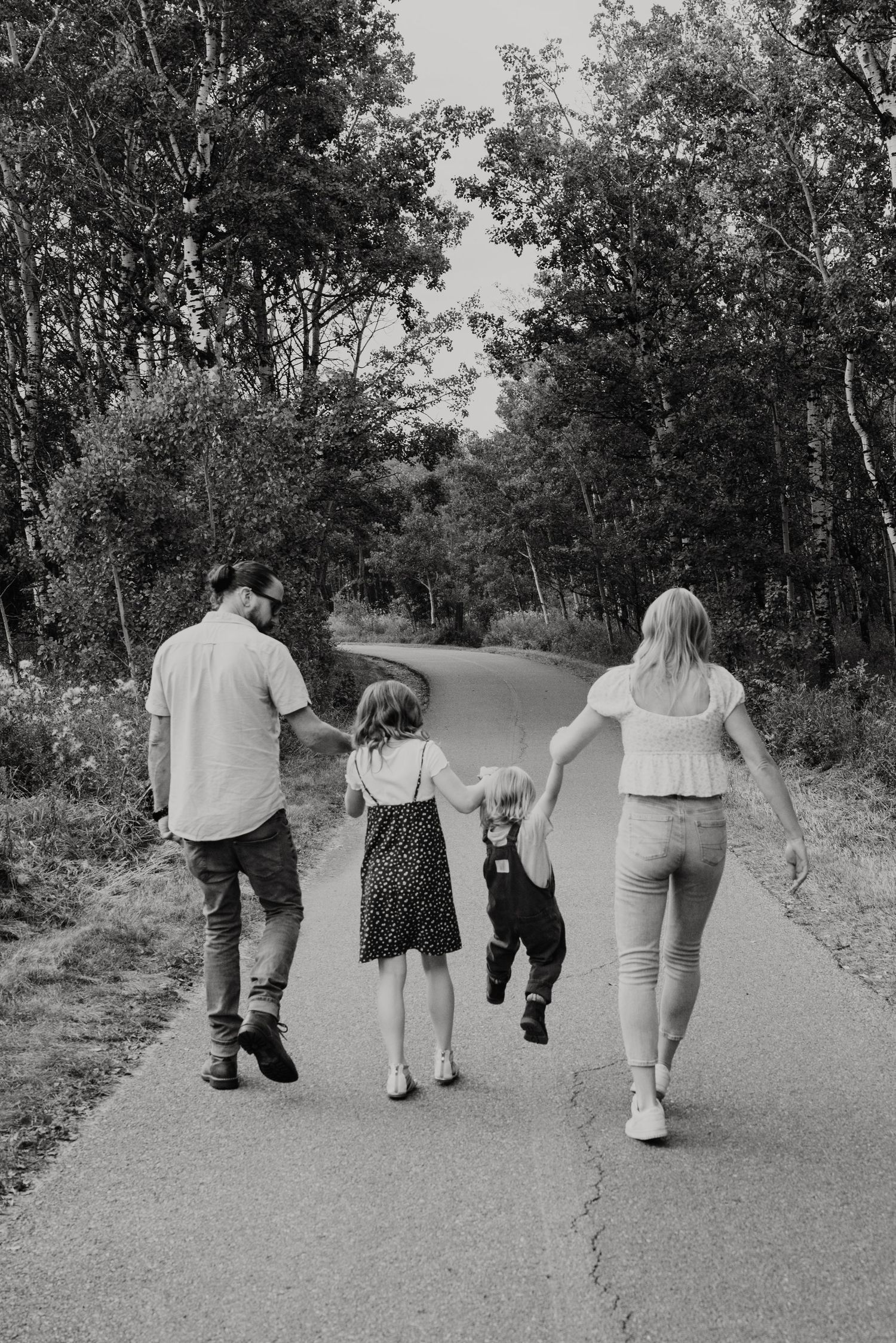 Black and white photo of a family of four walking hand-in-hand down a winding tree-lined path.