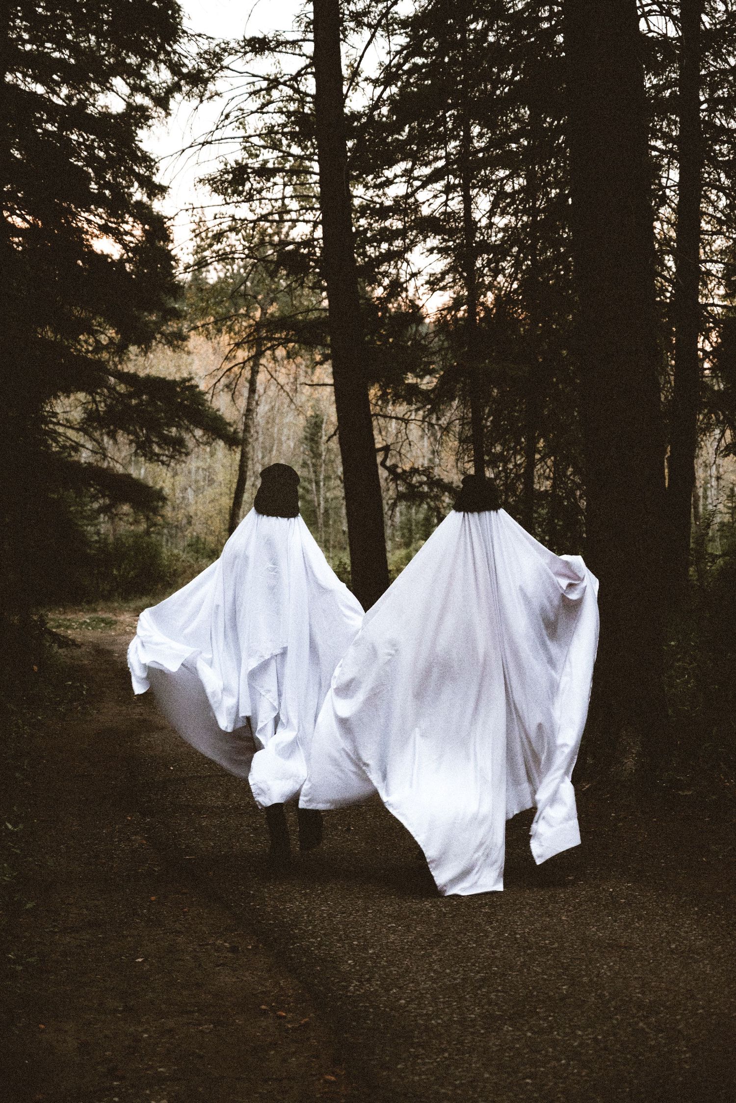 Two people in white ghost costumes walk along a forest path at dusk, sheets billowing behind them.