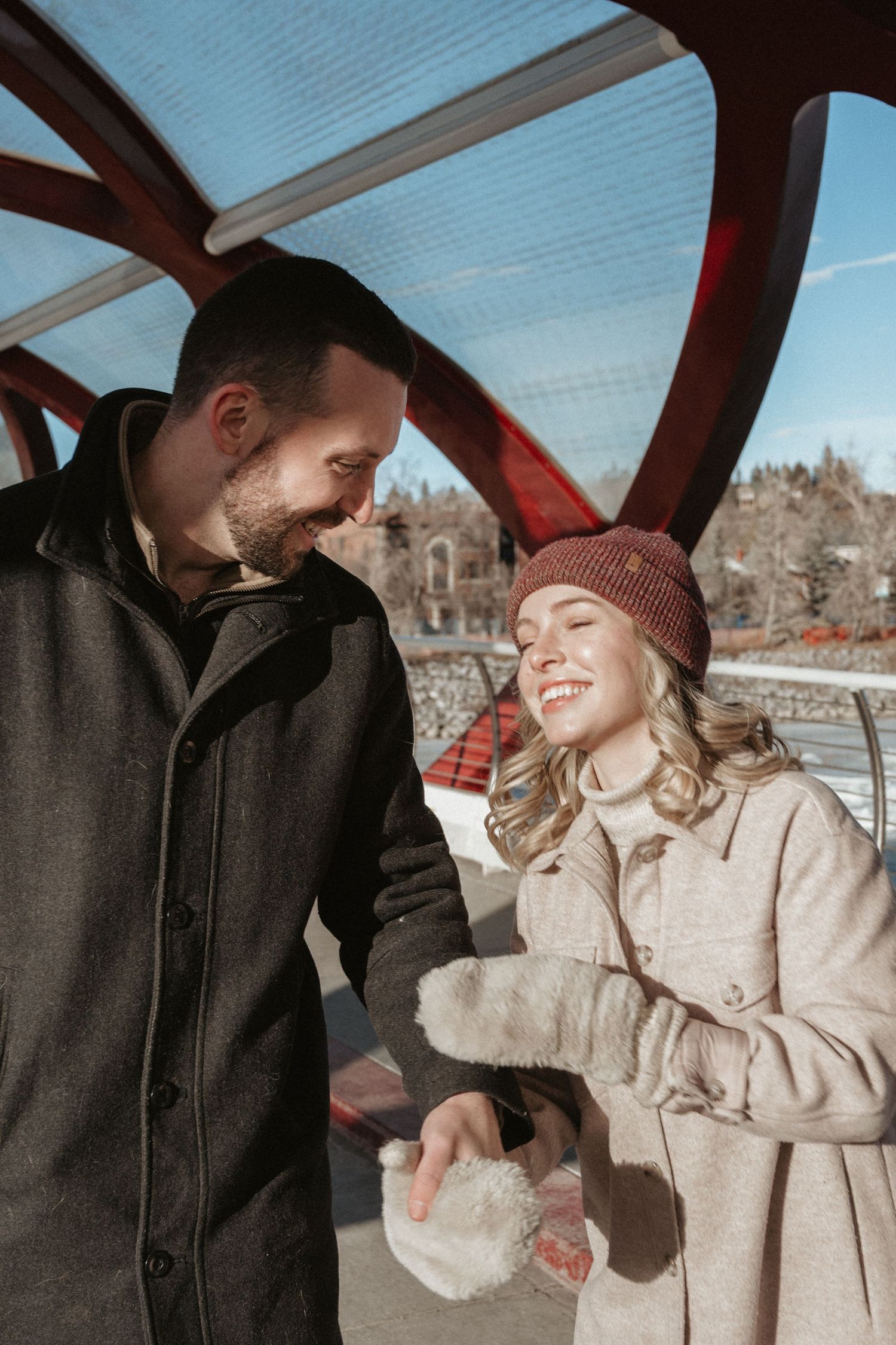 A happy couple in winter coats smiling at each other under a red arch bridge on a sunny day.