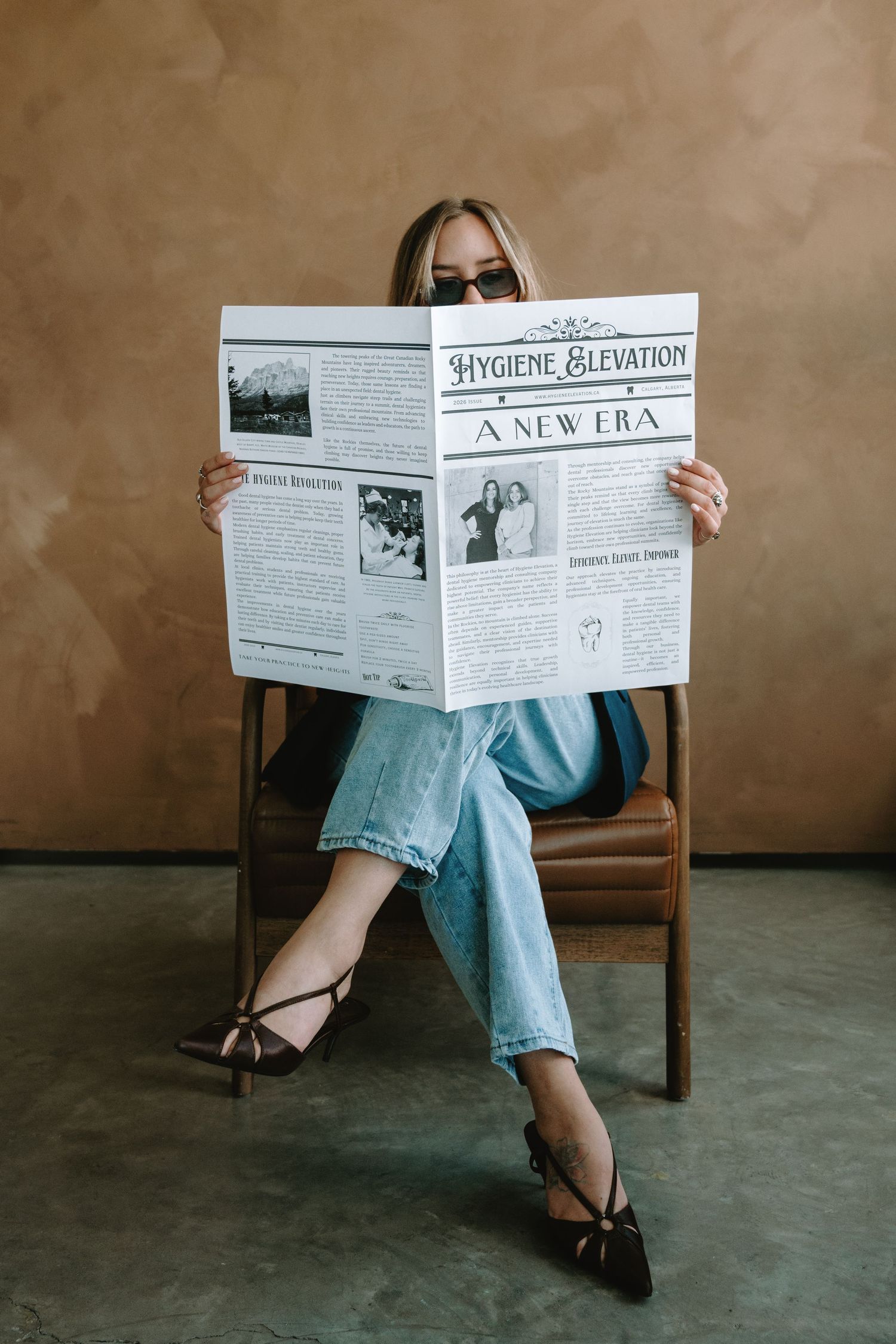 Woman sitting in chair reading Hygiene Elevation newspaper, wearing jeans and sunglasses in stylish editorial setting.