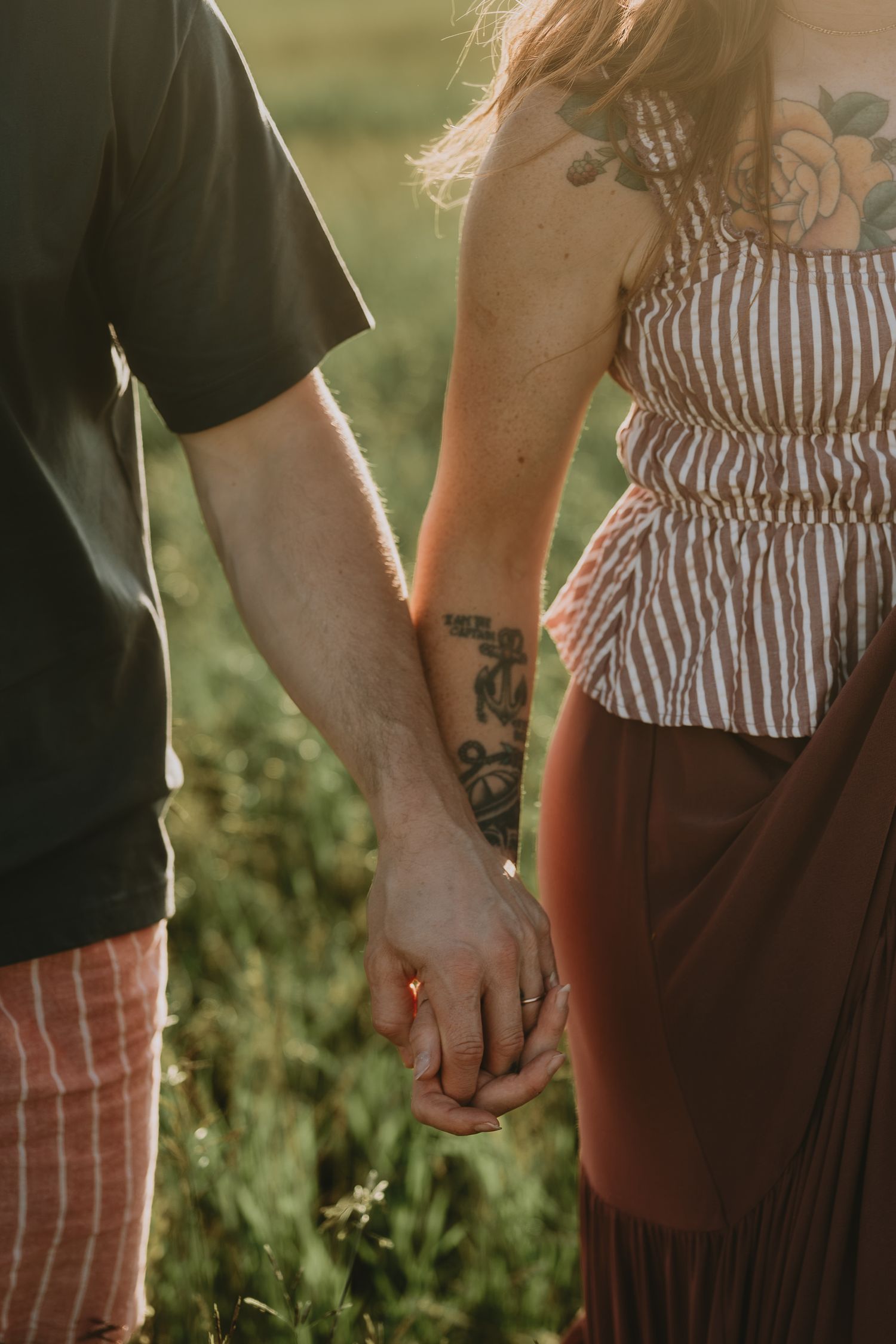 Couple holding hands with tattoos visible, standing in a sunlit field, wearing casual summer outfits.