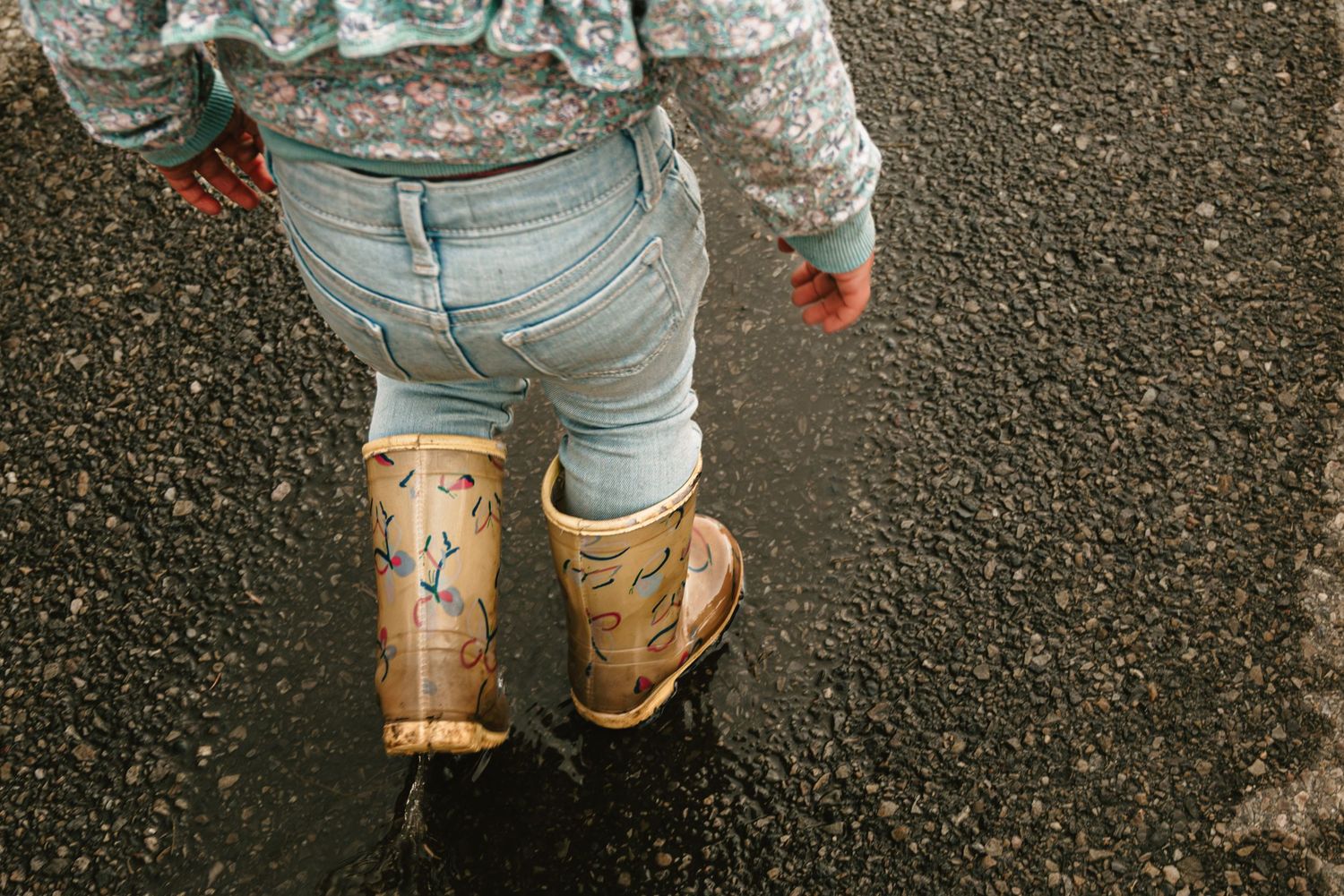 A toddler in jeans and decorative rain boots stands in a puddle on a rainy day.