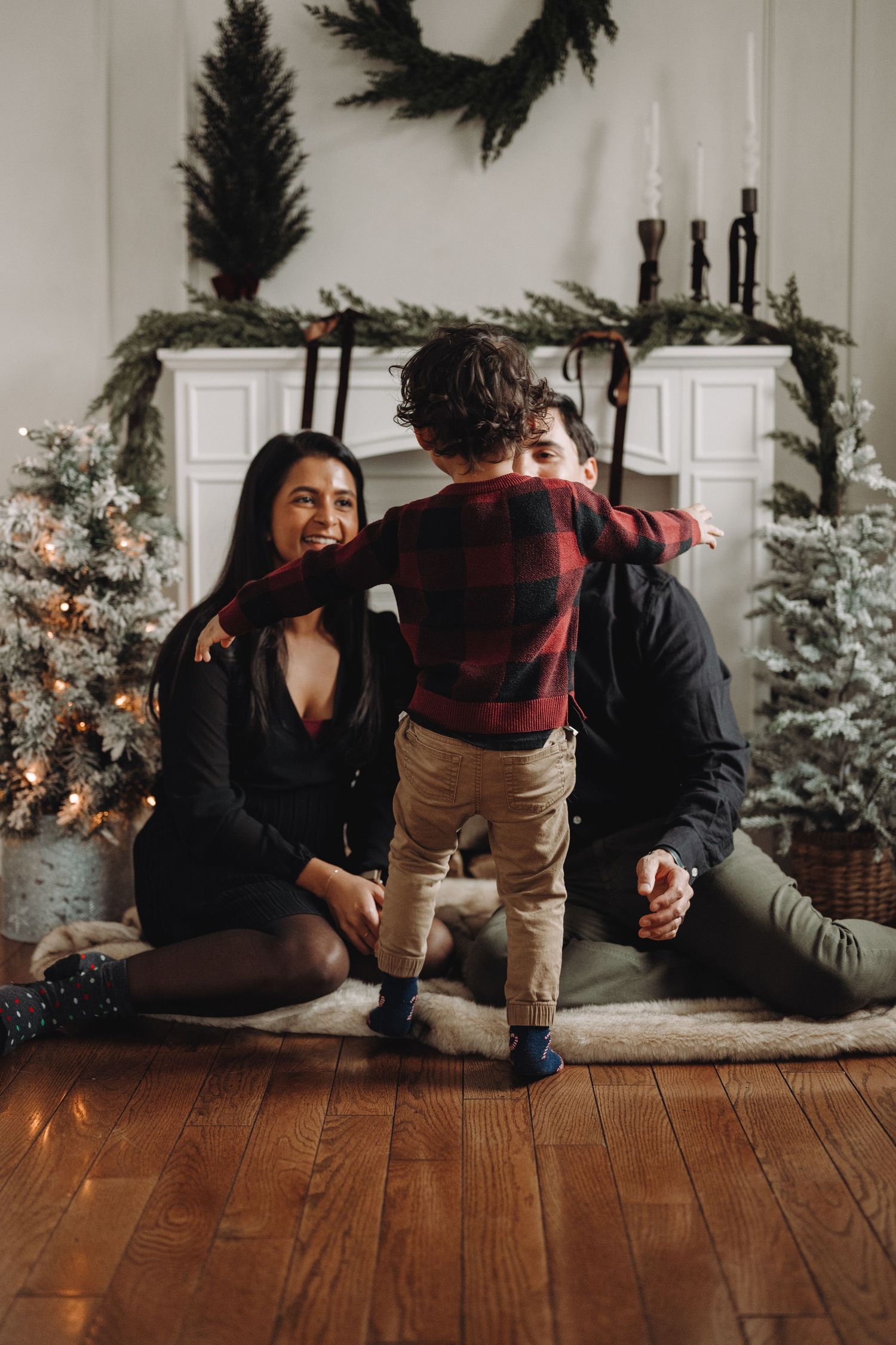 Family celebrating Christmas with a young child playing between parents on a hardwood floor near decorated trees and fireplace.