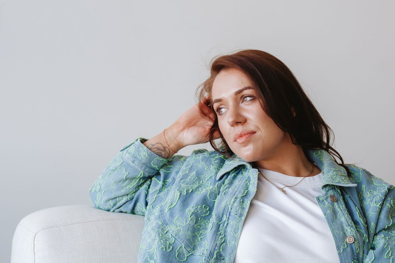 Young woman in floral green jacket sitting on white sofa, gazing thoughtfully to the side.