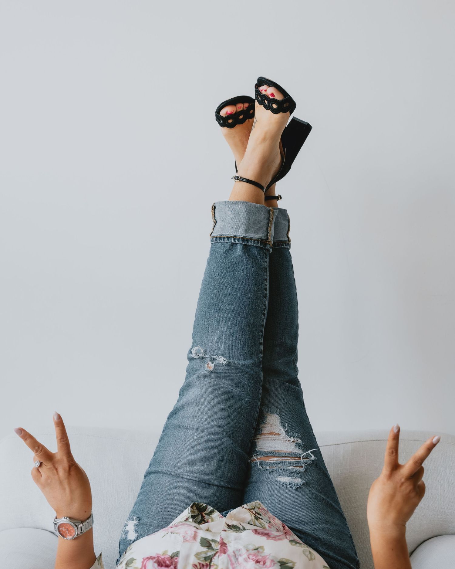 Woman in ripped jeans with black heeled sandals raised up, making peace signs on sofa.
