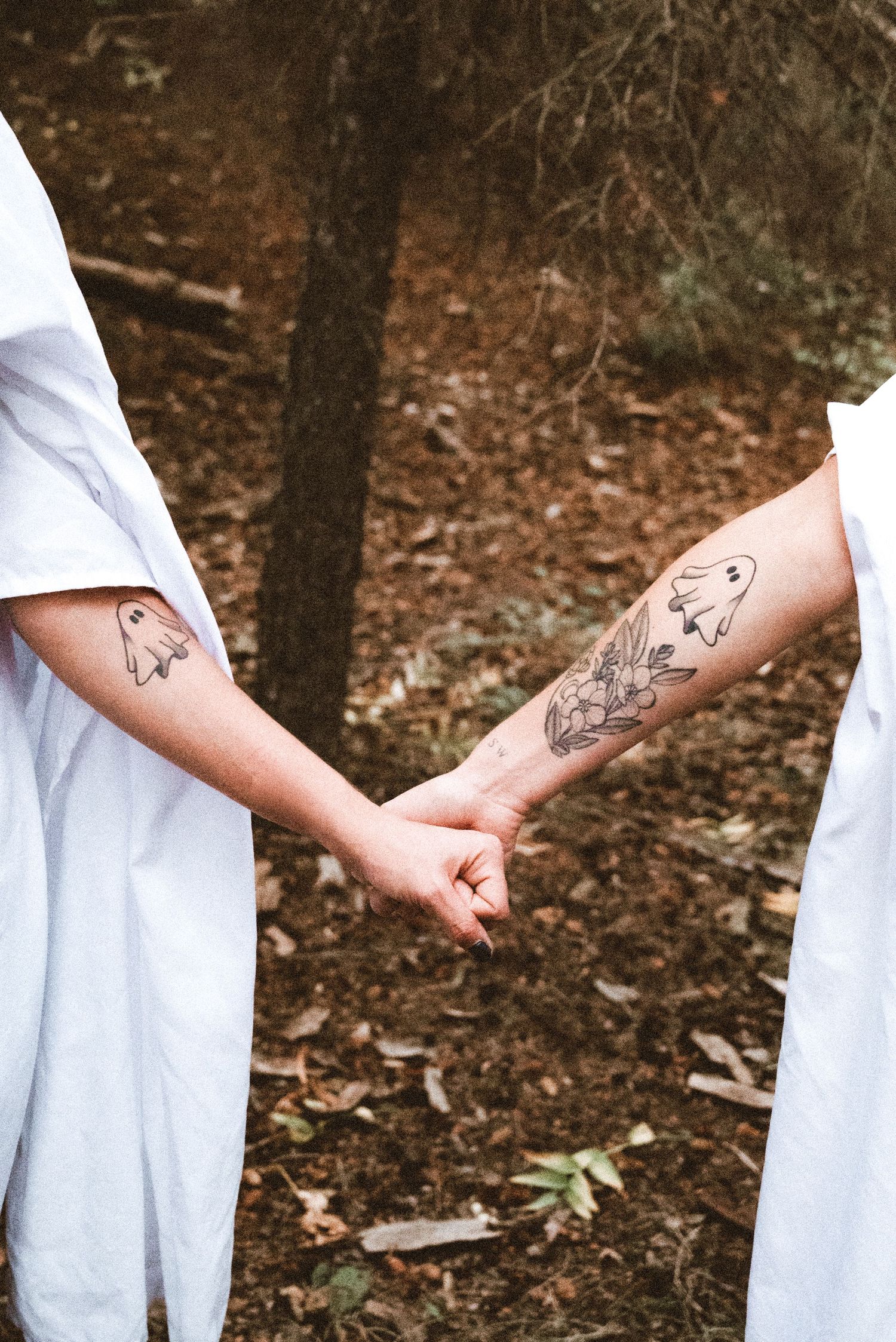 Two people holding hands in a forest, both wearing white, with matching ghost tattoos on their forearms.