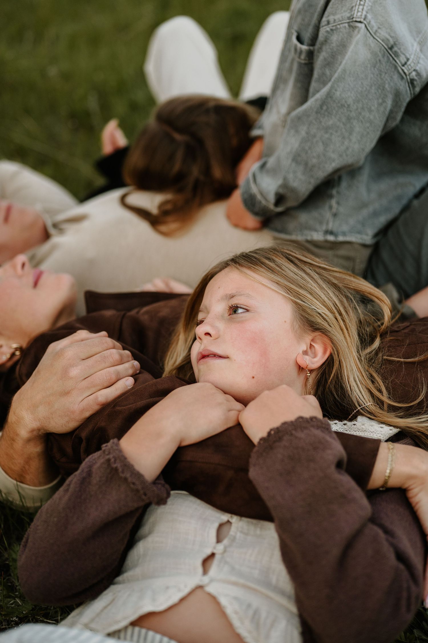 A young girl lying on grass with family members, looking up thoughtfully during an outdoor portrait session.