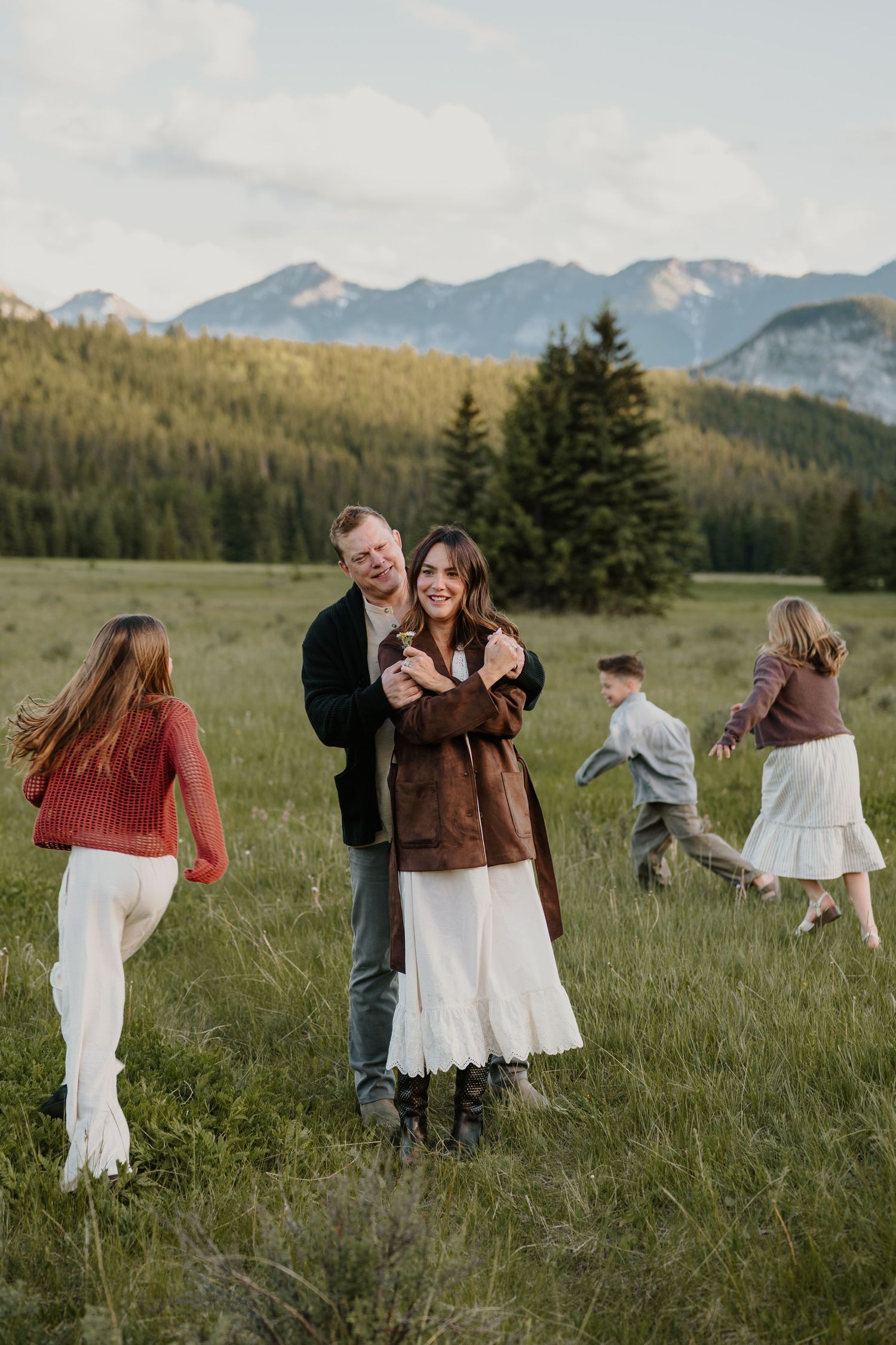 Smiling couple embracing in mountain meadow in Banff at golden hour while children play in background.