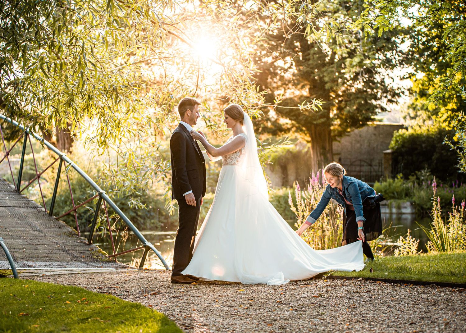 Bride and groom share a romantic moment in a sunlit garden while an assistant arranges the wedding dress train.