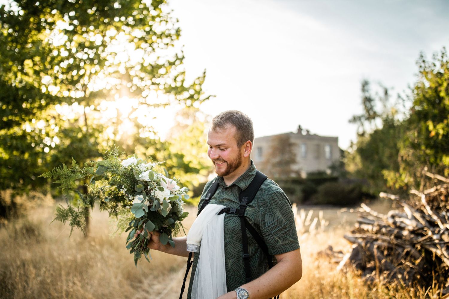 Bearded groom holding white bridal bouquet in golden sunlight at rustic outdoor wedding location.