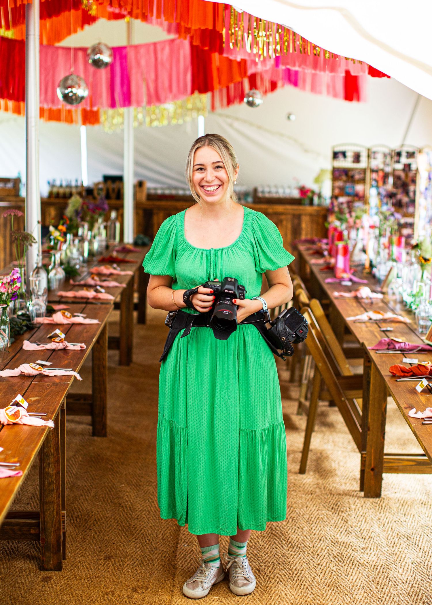 Wedding photographer in a green dress holds her camera while standing in a colorfully decorated reception venue.