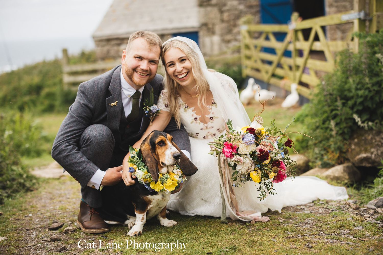 Bride and groom smile with their beagle wearing a floral collar outside a rustic stone building at their wedding.
