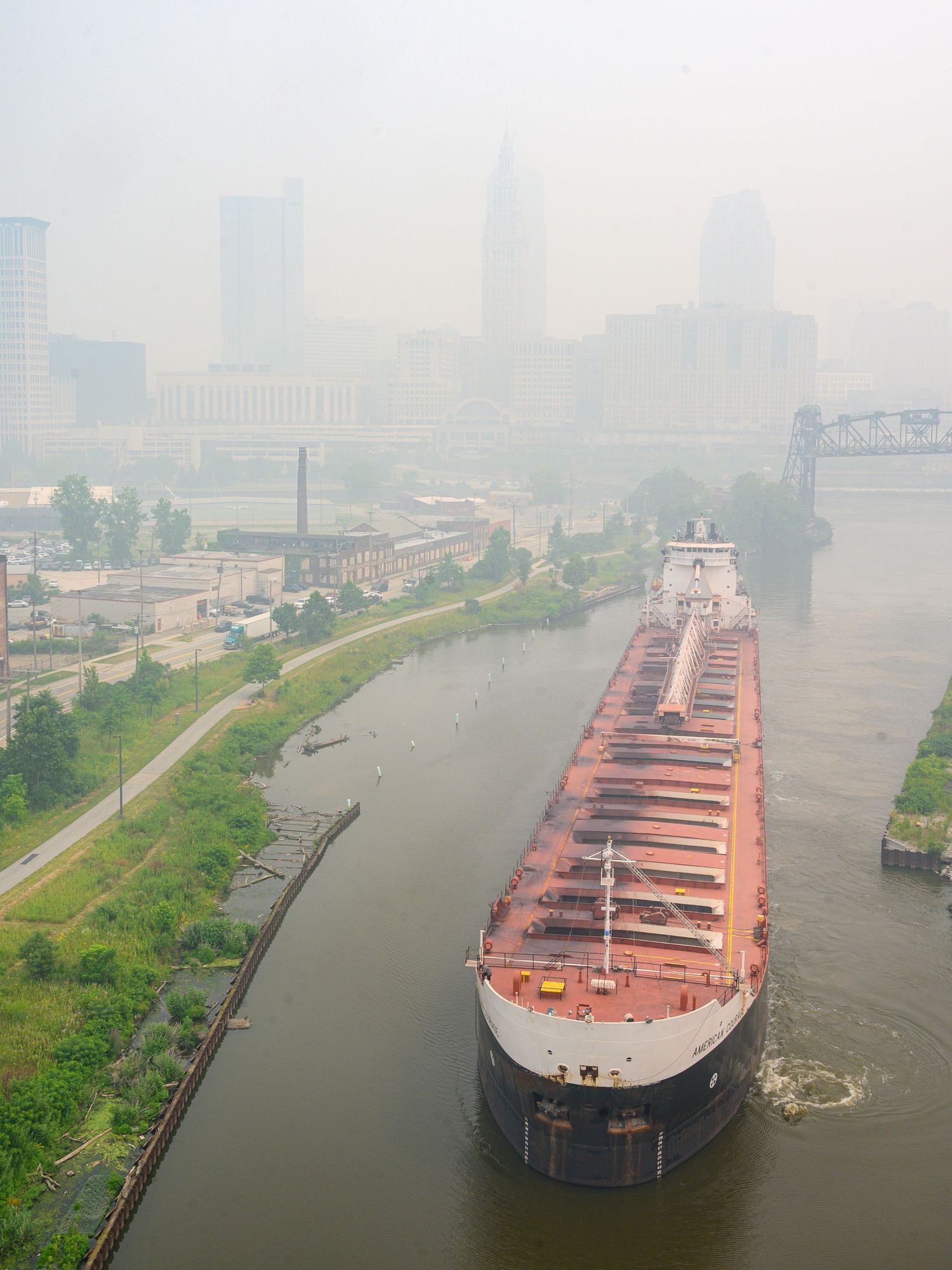Large cargo ship navigating foggy river through industrial city waterway on hazy day.