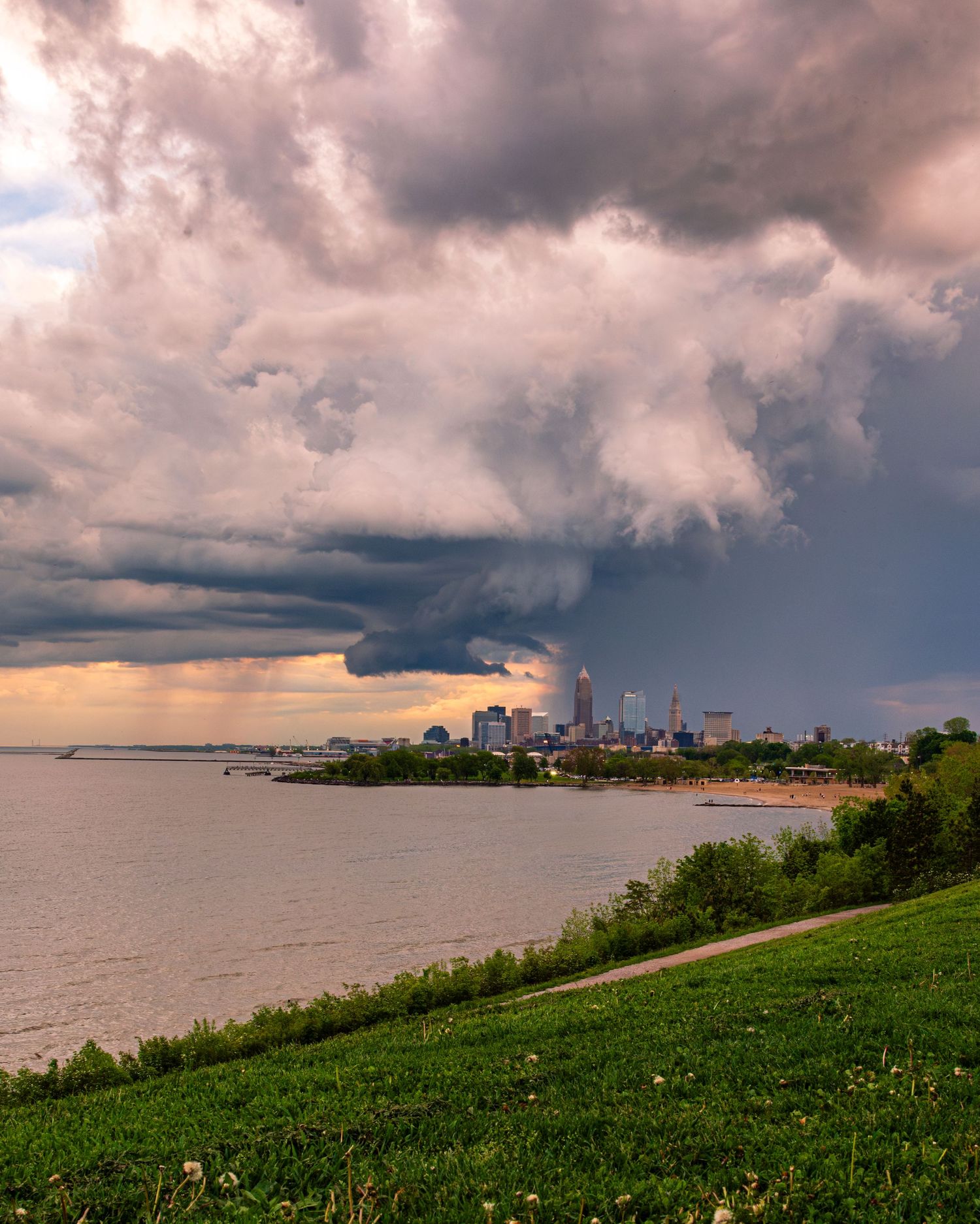 Storm clouds gather over Cleveland skyline viewed from a grassy lakefront park.
