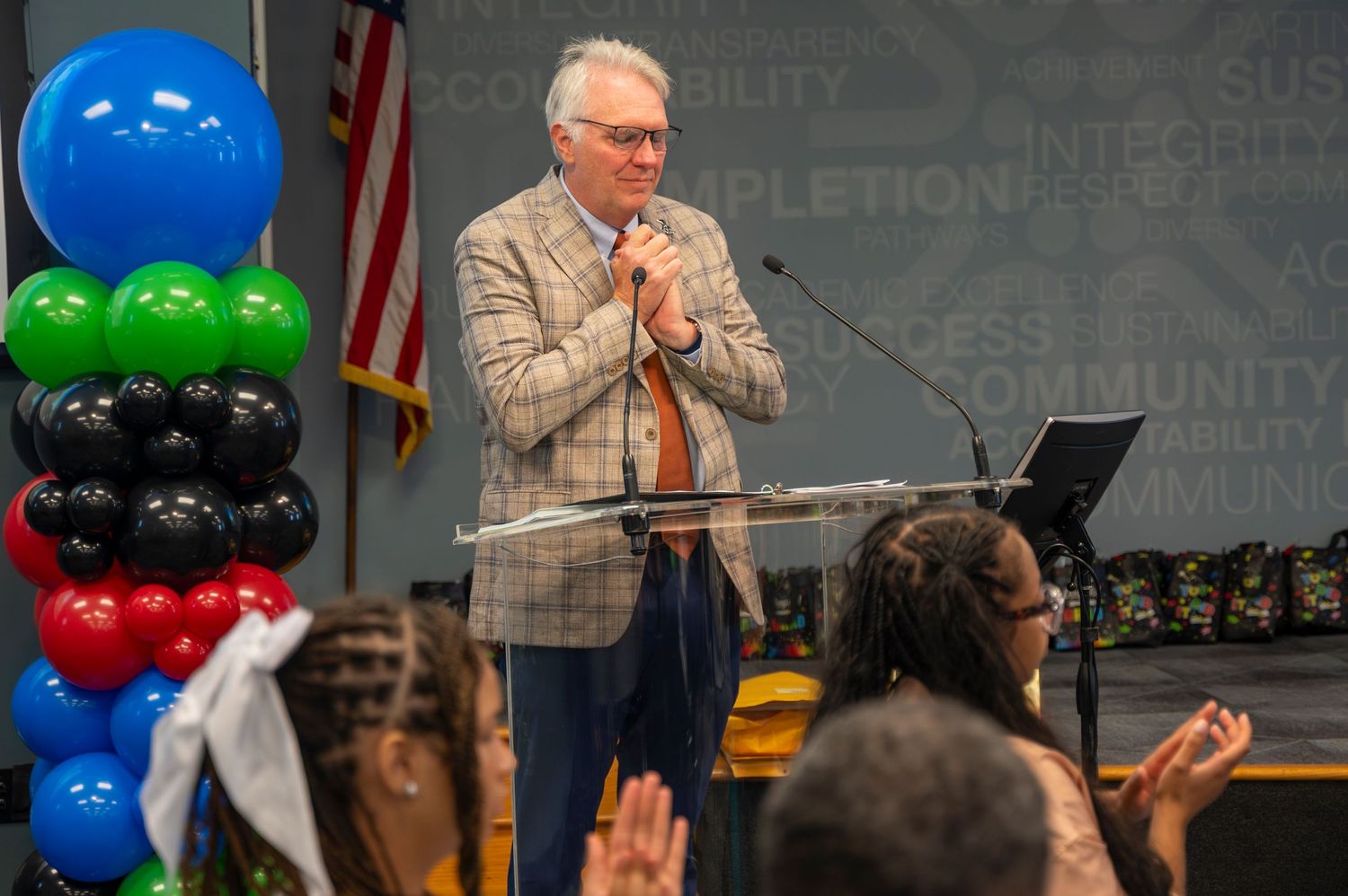 Speaker in plaid blazer gestures to audience at graduation ceremony podium with balloons.
