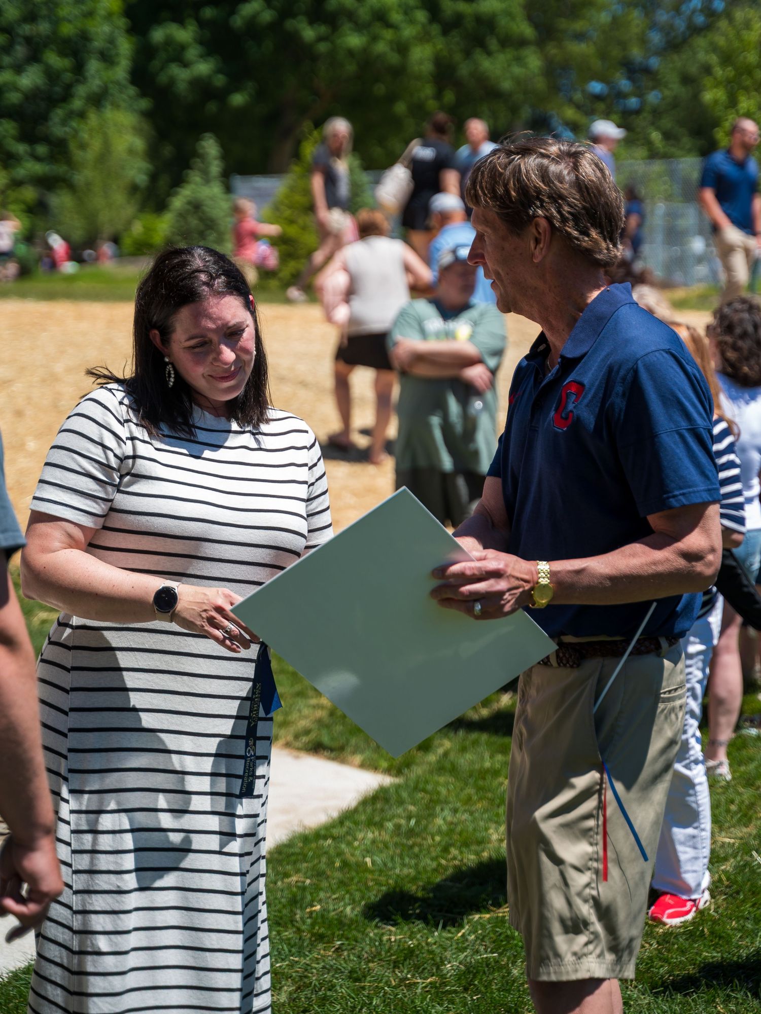 Two people conversing outdoors at a sunny event, woman in striped dress and man in blue polo holding a green folder.