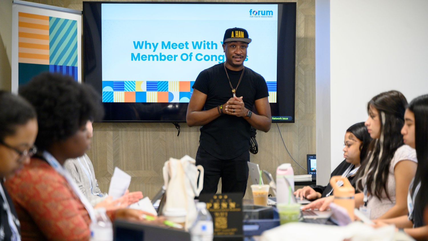 A man presents to a diverse group of students in a modern classroom with a screen showing Why Meet With a Member of Congress.