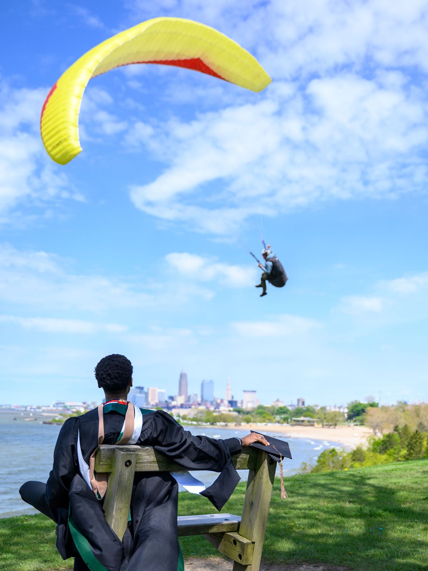 Person sitting on a bench watching a paraglider soar over a scenic lakeside city skyline.