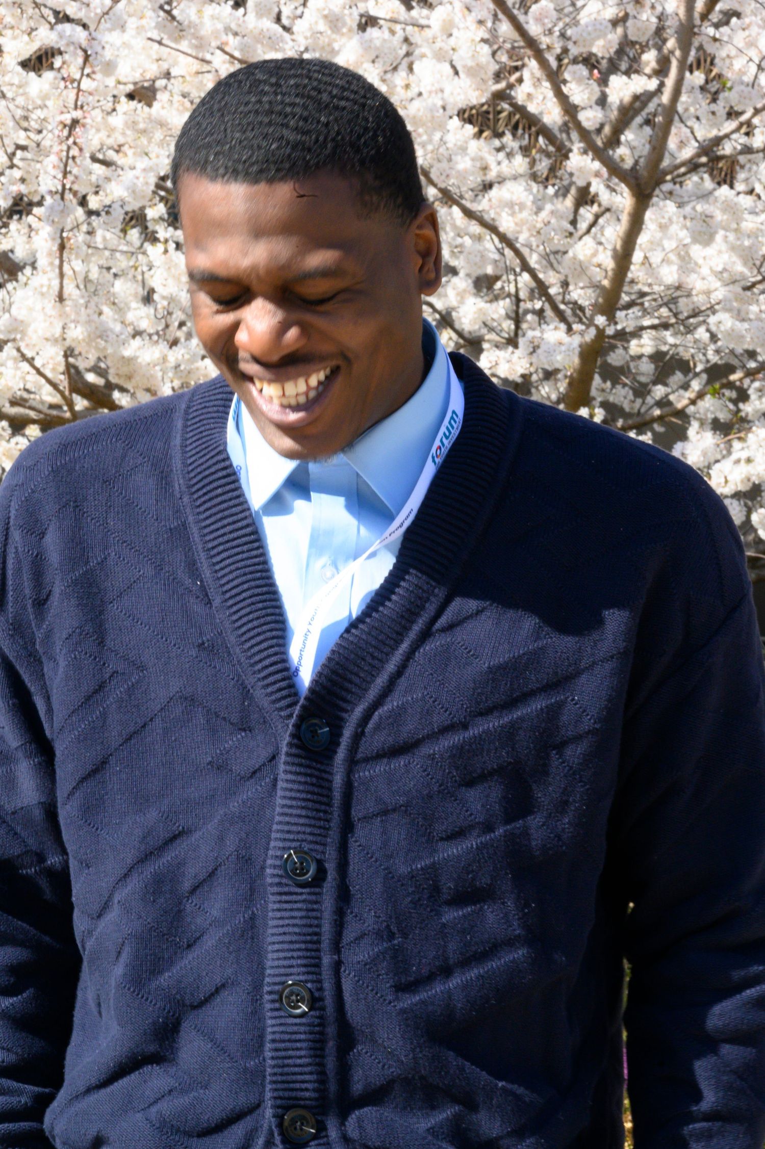 Smiling young man in navy cardigan standing in front of blooming cherry blossom trees.