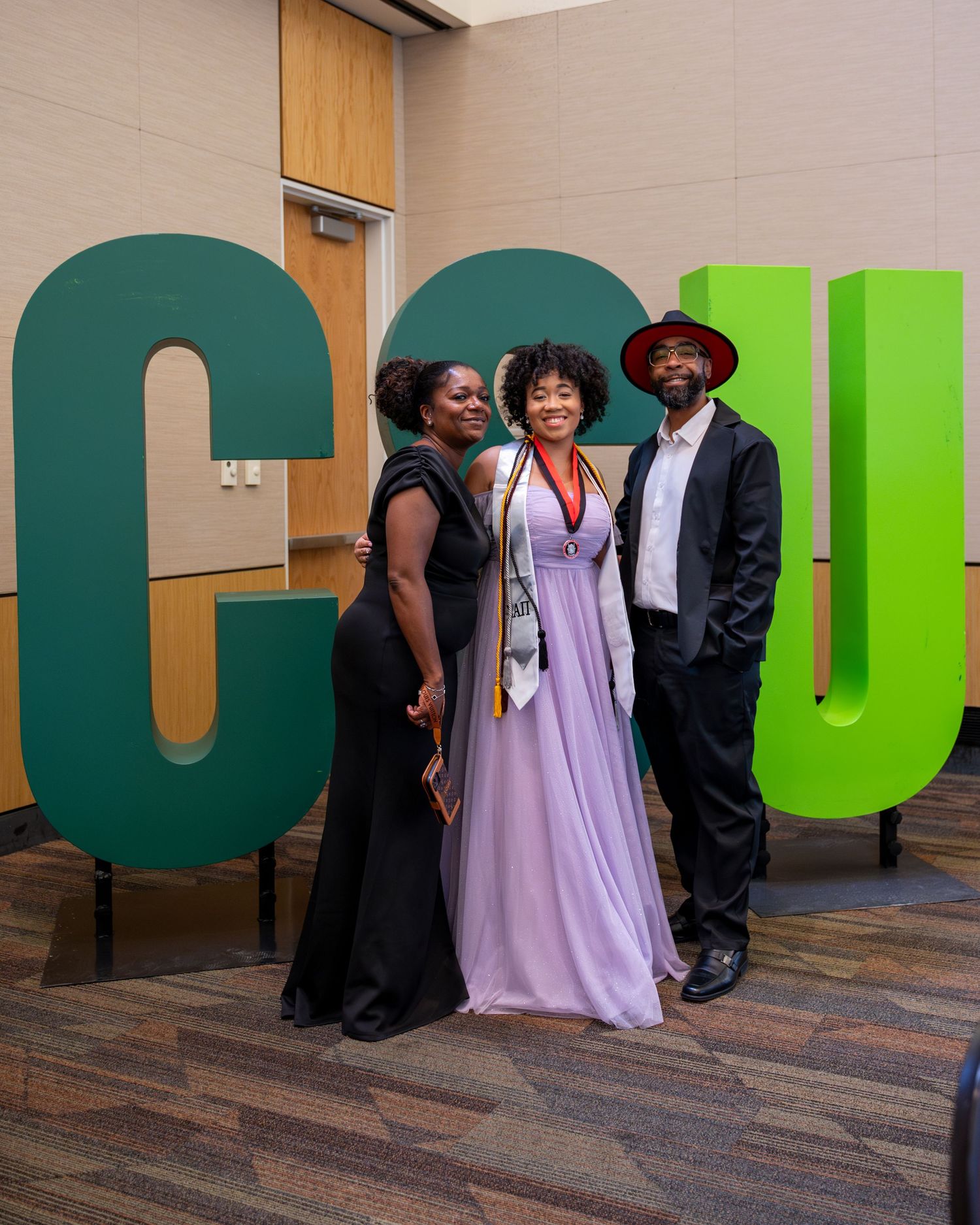 Three people posing in formal attire in front of large green CSU letter signs at a graduation event.
