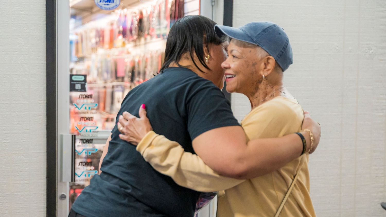 Two women share a warm hug near a store refrigerator display case.