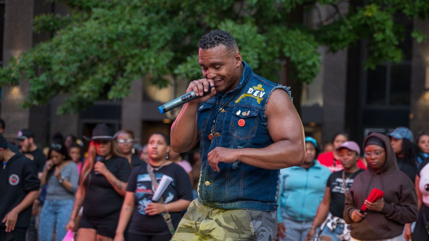 A muscular performer singing into a microphone at an outdoor concert with a crowd behind him.