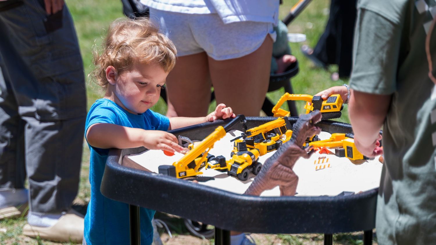 Young boy in blue shirt playing with yellow toy construction vehicles on an outdoor table at a sunny event.
